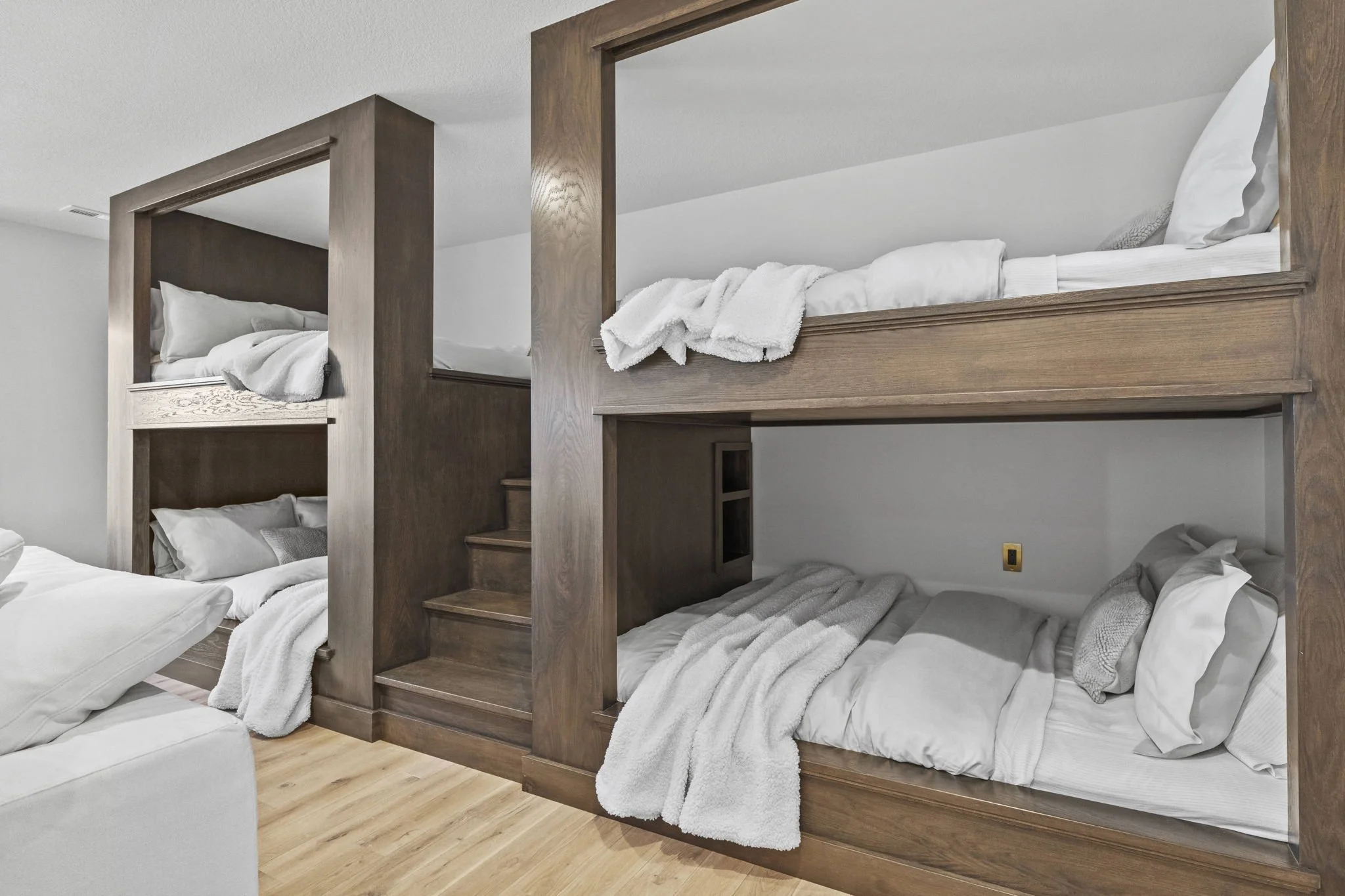 A set of triple bunk beds with wooden frames and stairs, with white bedding and pillows, in a room with light-colored walls and wooden flooring.