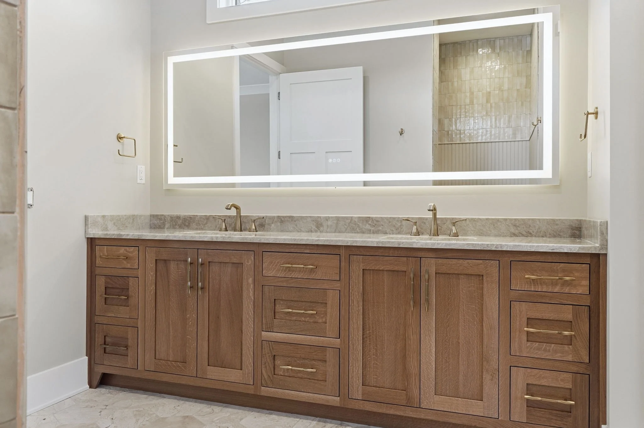 Bathroom vanity with wooden cabinets, a marble countertop with two sinks, and a large backlit mirror.