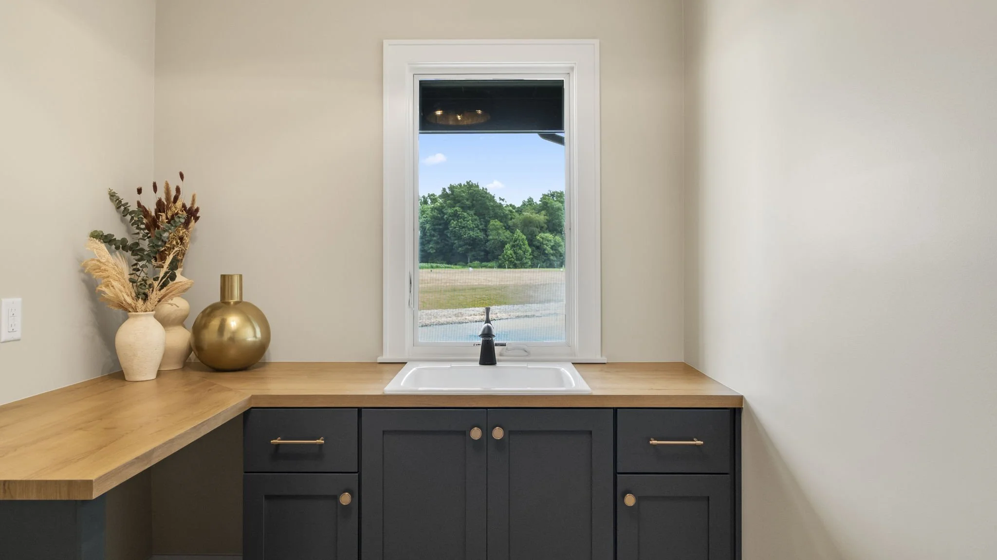A kitchen corner with beige walls, black cabinets, a wooden countertop, a white sink under a window, decorative vases with dried plants, and a view of greenery outside.