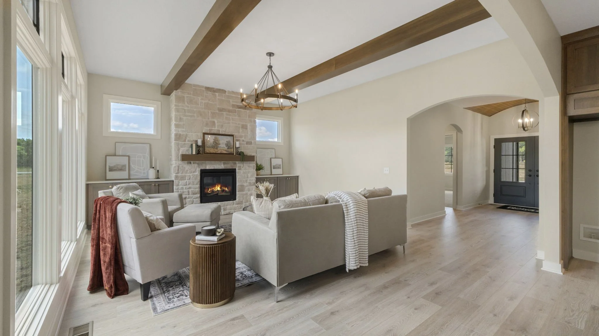Bright living room with large windows, white sofas, a stone fireplace, and wooden ceiling beams, decorated with framed art and plants.