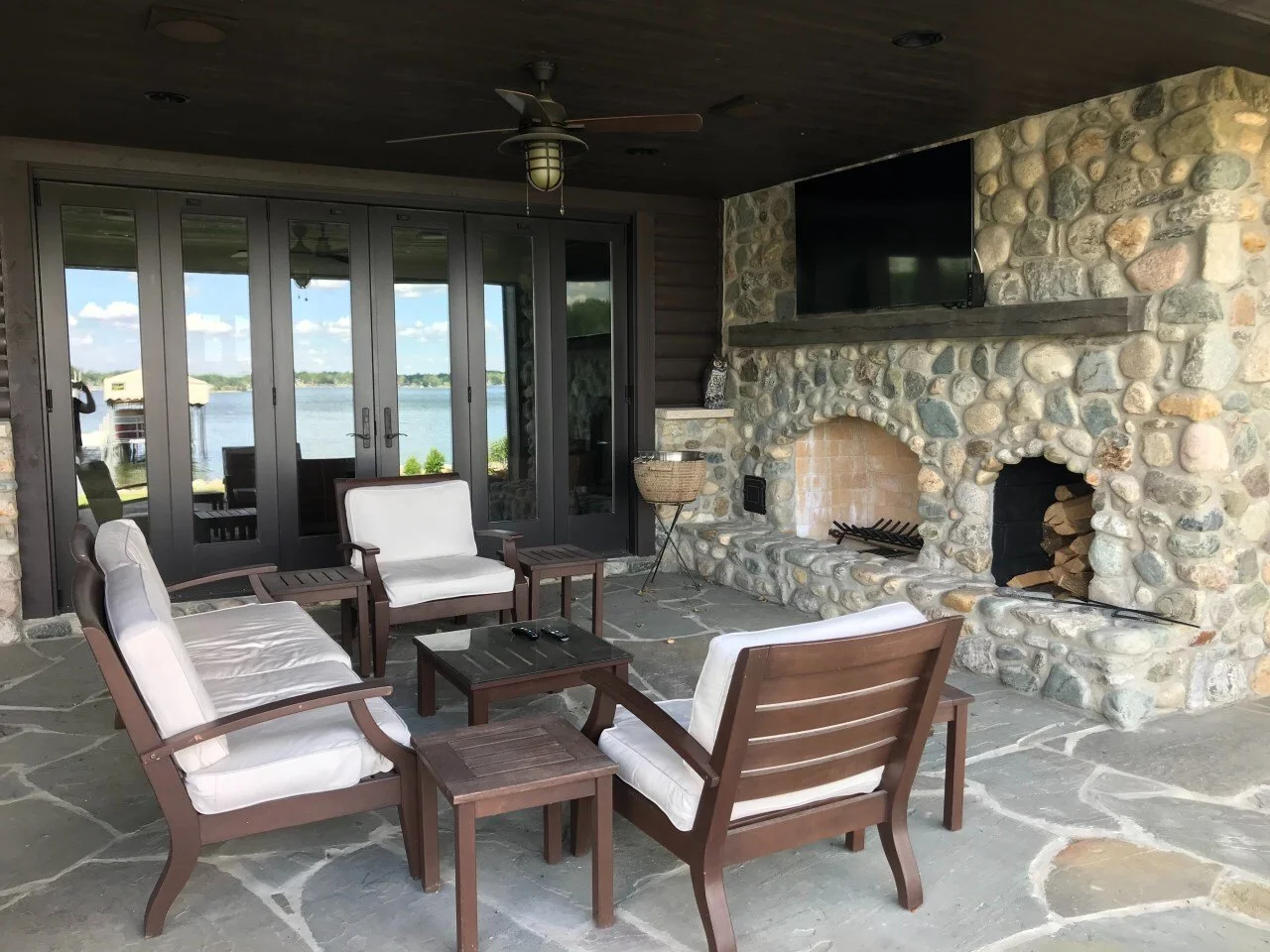 Outdoor patio with wooden furniture, ceiling fan, stone fireplace, flat-screen TV, and glass doors overlooking a lake.
