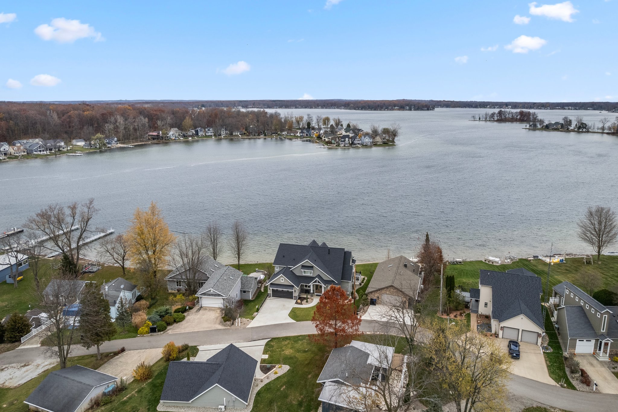 Aerial view of lakeside houses along a shoreline, with a large water body in the background and a clear blue sky with scattered clouds.