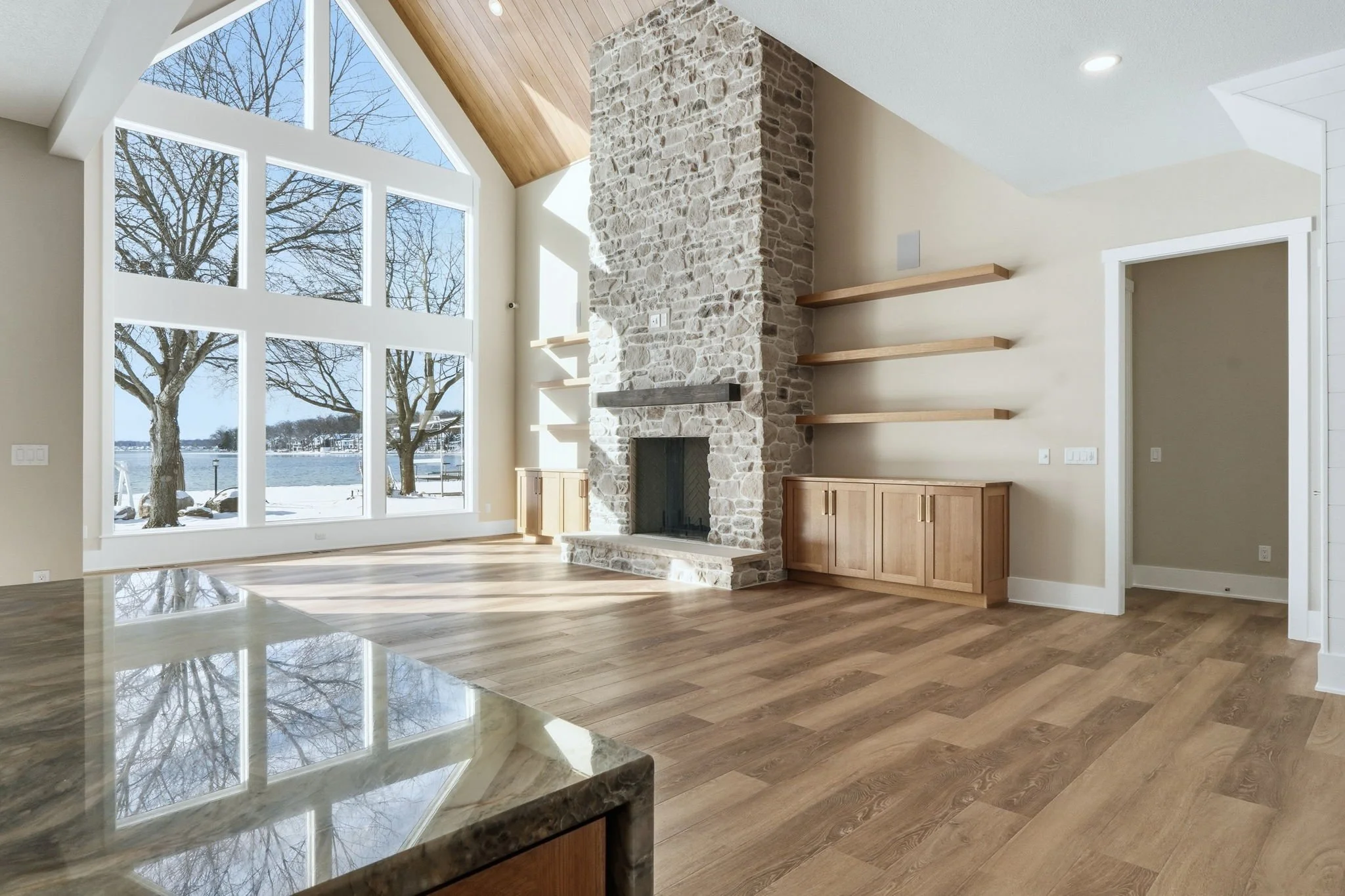 Bright living room with large floor-to-ceiling windows overlooking a snowy outdoor landscape, a stone fireplace, floating wooden shelves, and hardwood flooring.