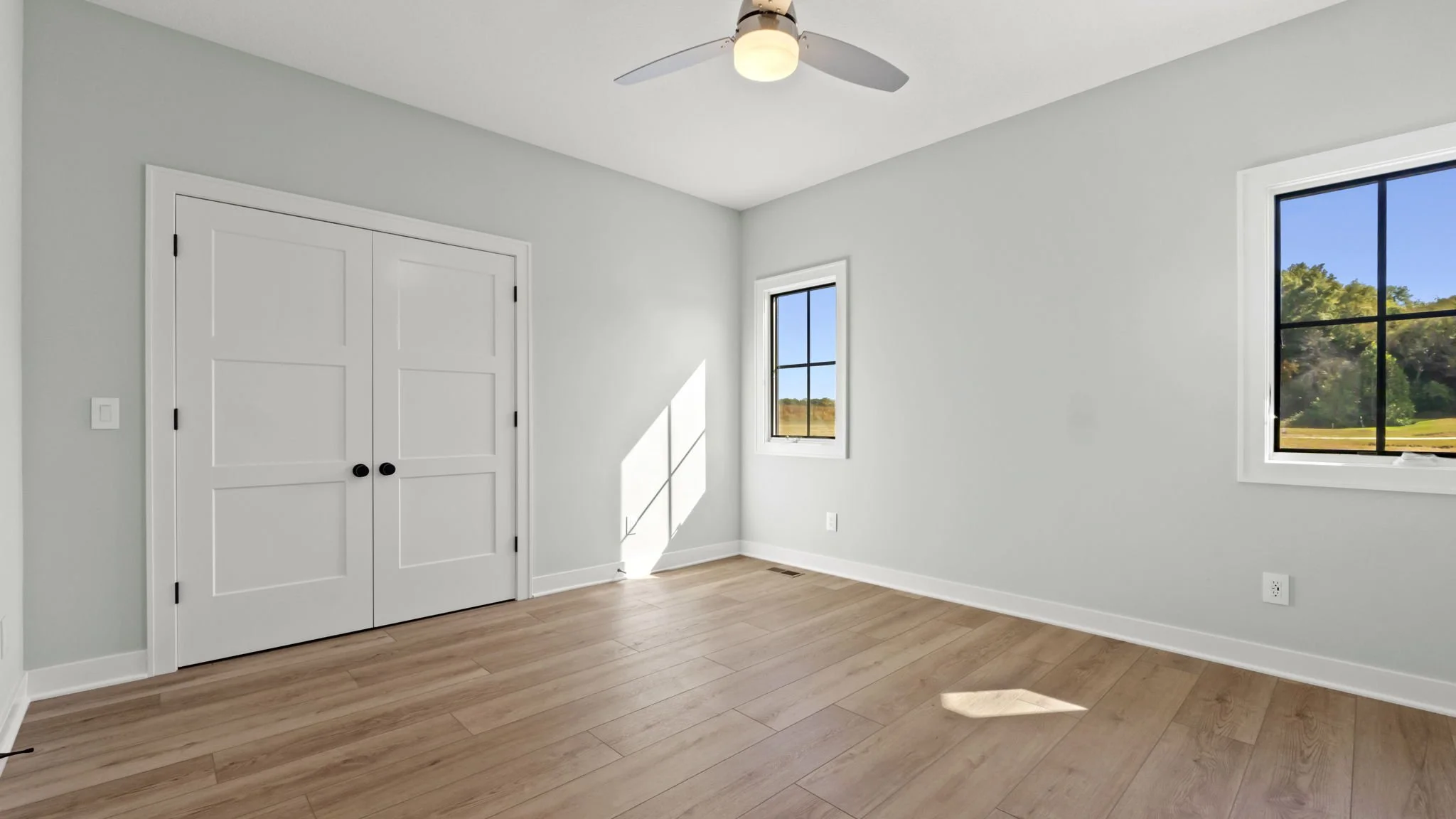 Empty bedroom with light gray walls, wood flooring, two windows showing outdoor scenery, a white double closet door, a ceiling fan with light, and electrical outlets.