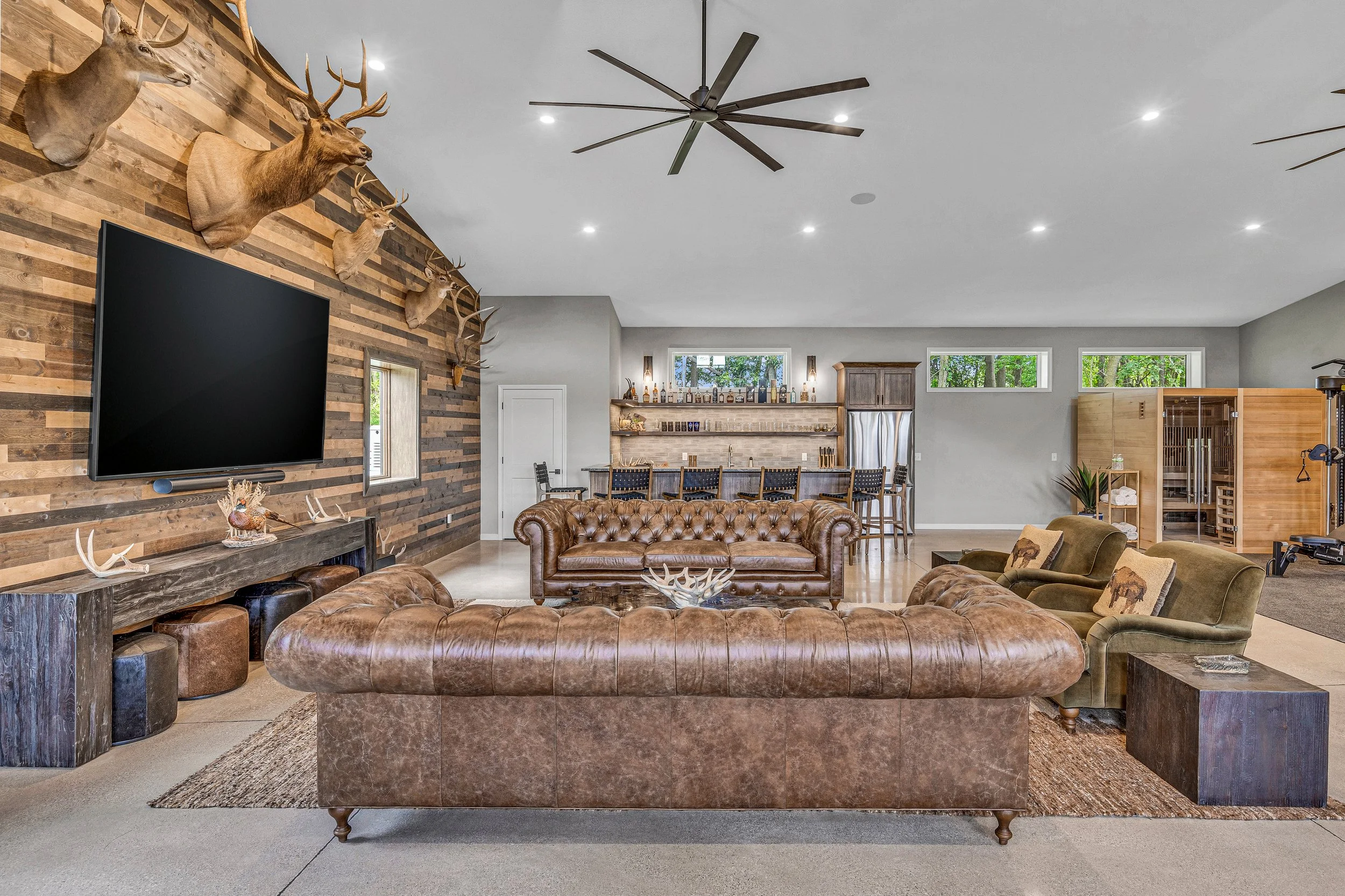 Living room with a mounted deer heads on a wooden wall, brown leather sofas, a large flat-screen TV, and a bar area with stools in the background. Deer-themed pillows on the side chairs. Rustic and modern decor with wood accents and festive lighting.