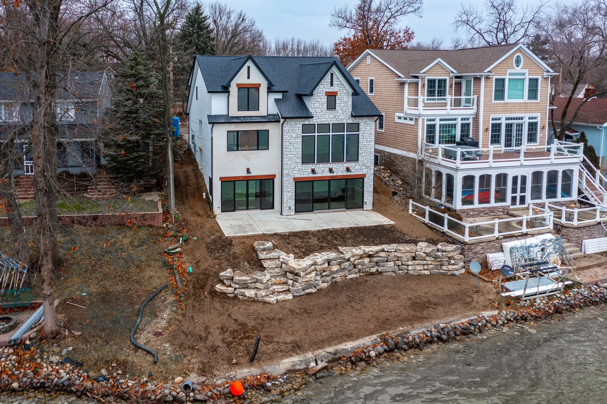 A partially constructed house with large windows on a sloped backyard by a body of water, featuring a stone retaining wall, concrete patio, and neighboring houses with decks and railings.