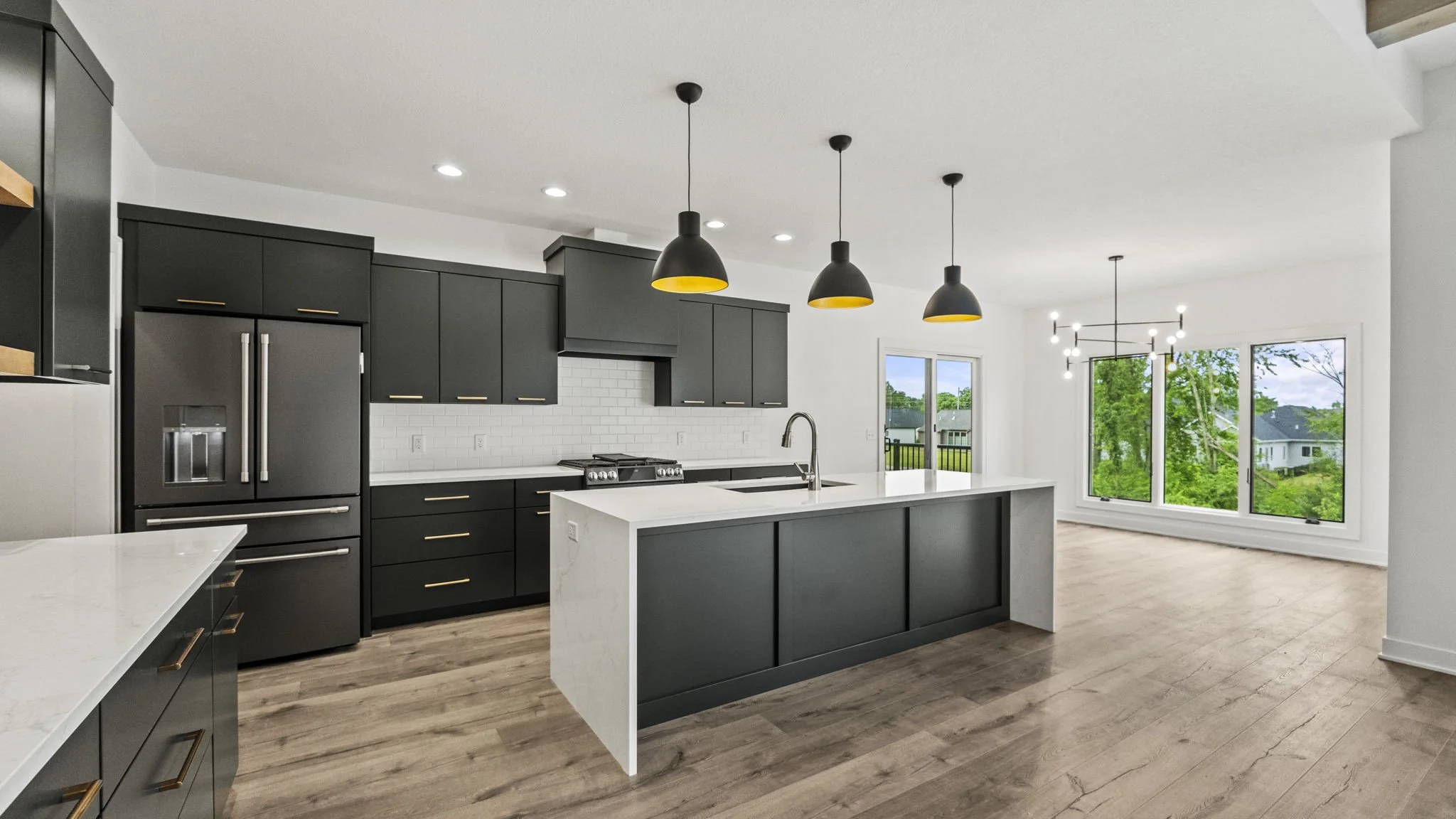 Modern kitchen with dark cabinets, white countertops, a large island, pendant lights, and large windows showing greenery outside.