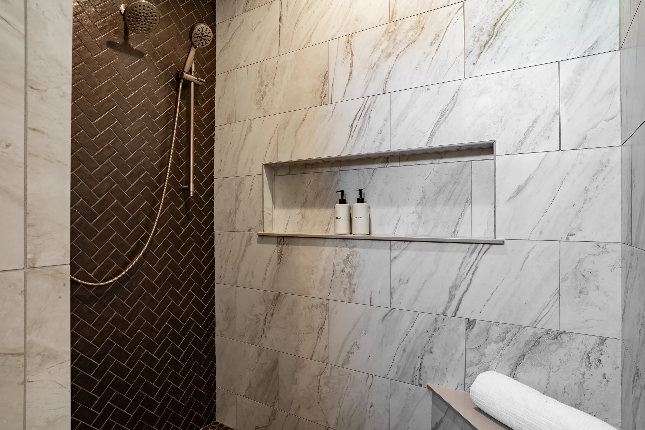 Modern shower area with dark herringbone tiles on the shower wall and light marble tiles on the main wall. Built-in shelf with soap and lotion bottles. White rolled towel on a shelf.