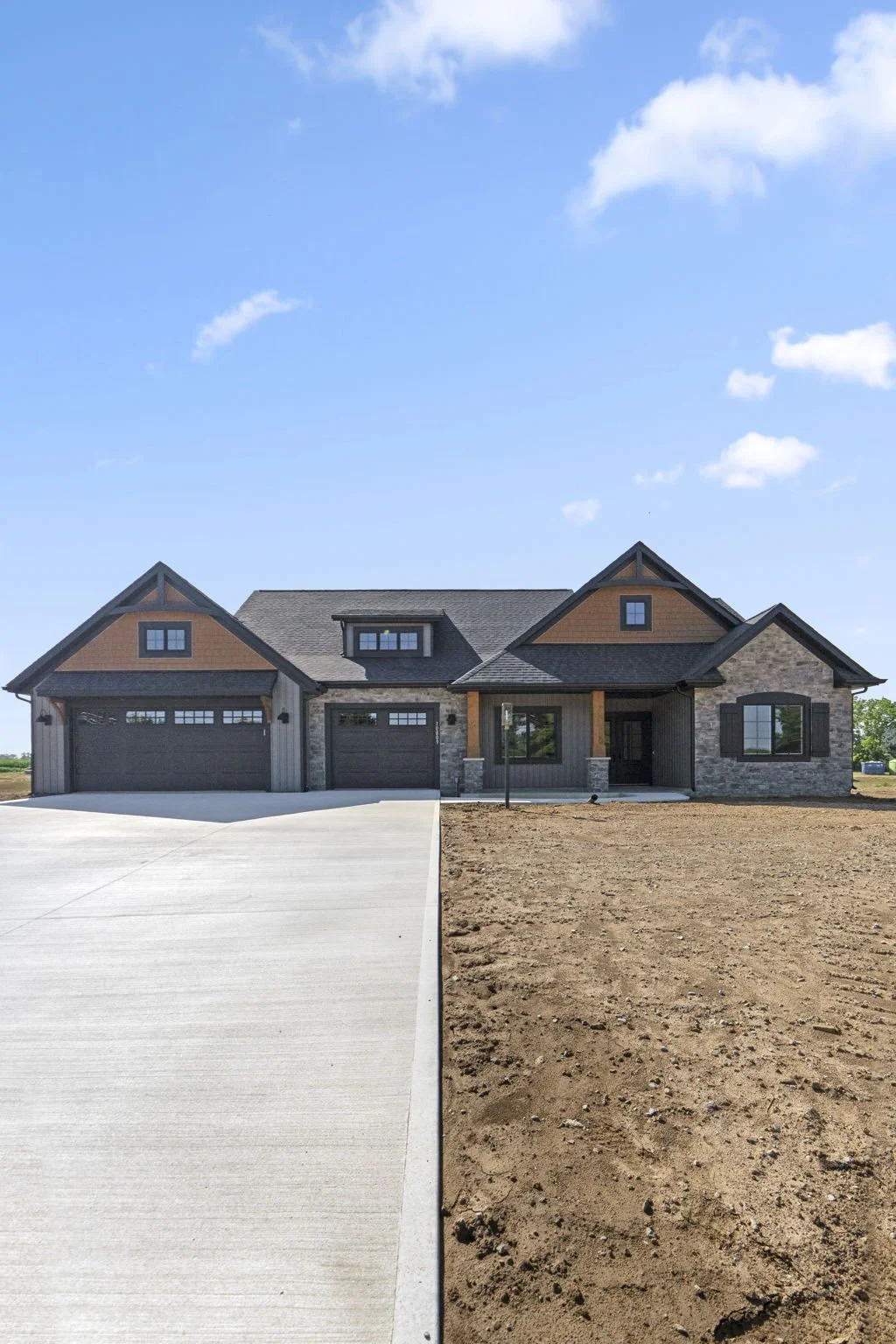 Newly built house with a two-car garage, brick and wooden exterior, and a front porch under a blue sky with clouds.