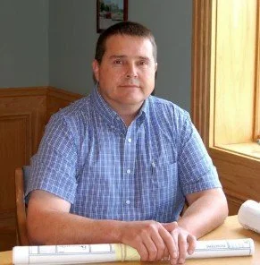 Man sitting at a desk with a blue checkered shirt, in front of a window with sunlight, holding a rolled-up paper.