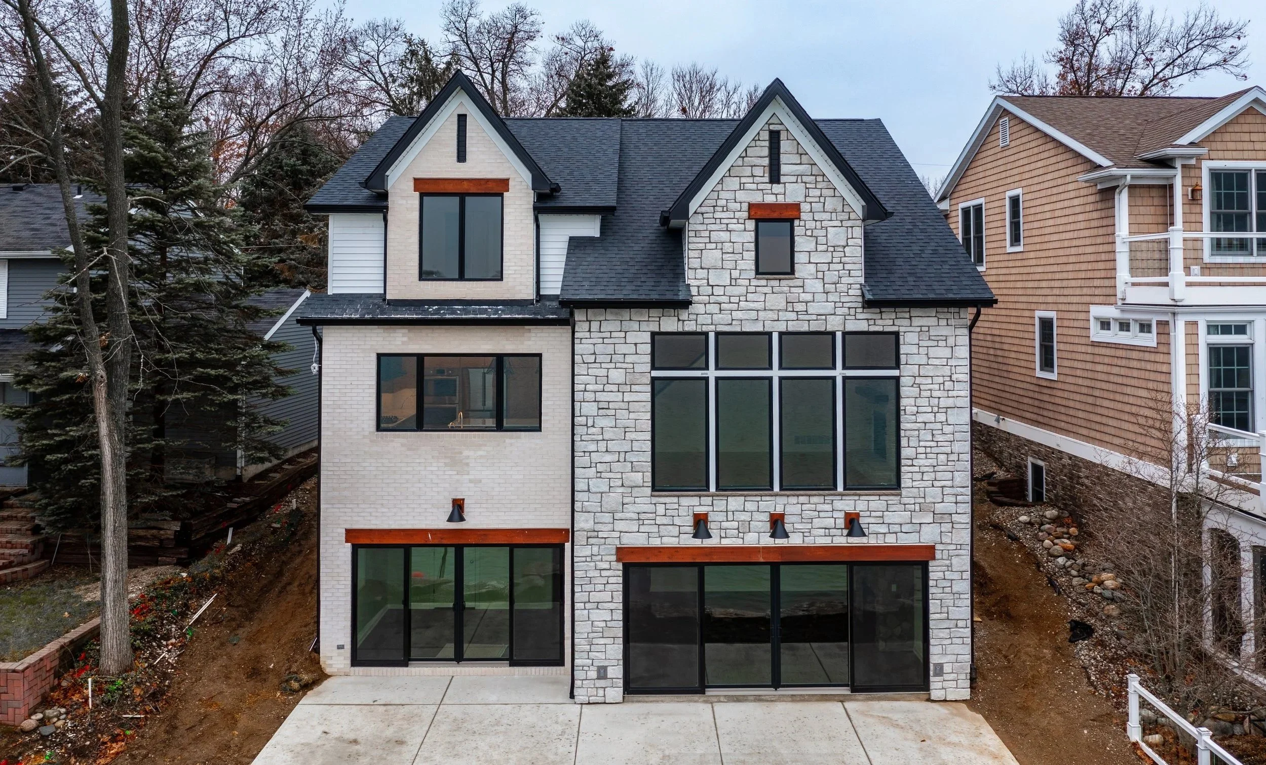 Newly constructed multi-story house with white brick and stone exterior, large windows, and a sloped roof, situated between two other homes, with a concrete driveway and surrounding landscaping.