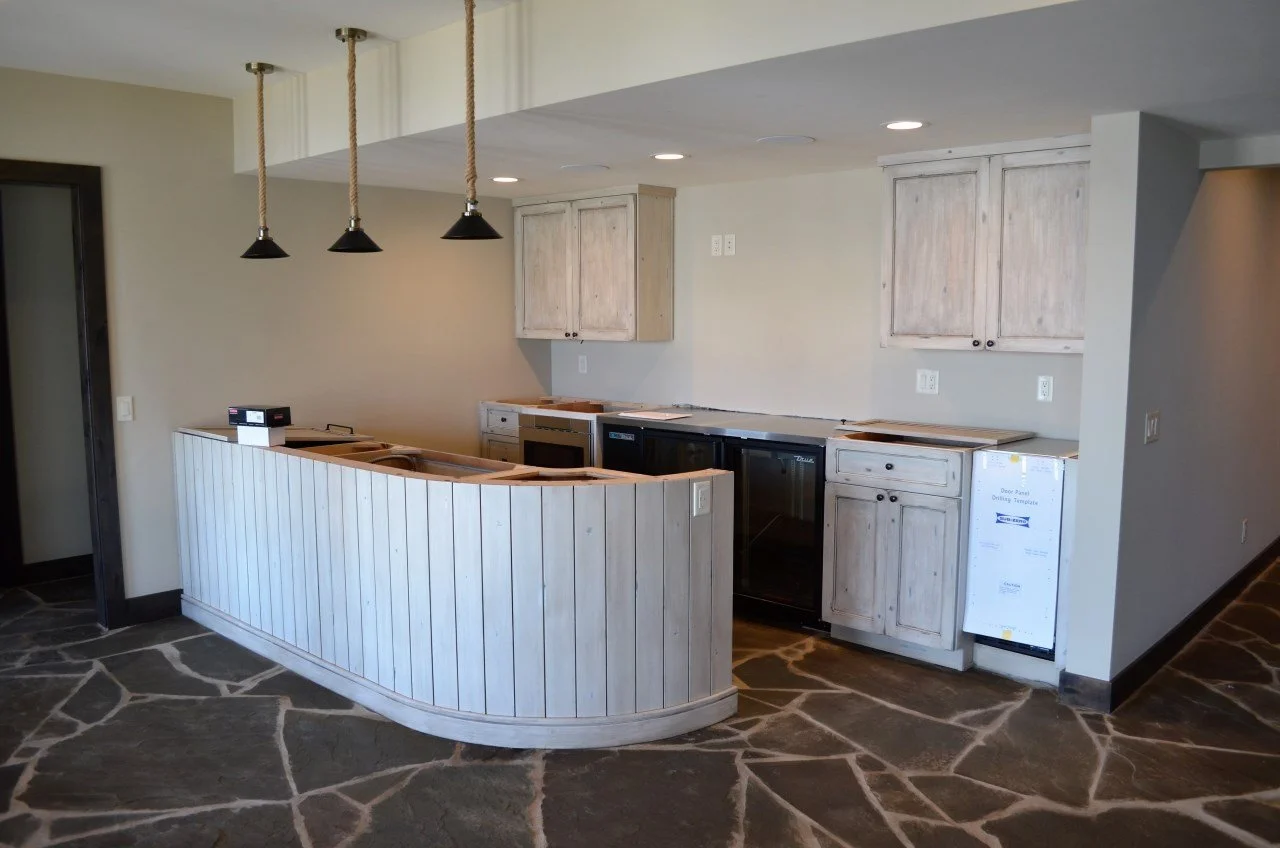 Kitchen area with white wooden cabinets, black appliances, and a curved bar with a light wood panel finish, featuring hanging lamps with black shades and rope accents, and a stone floor.