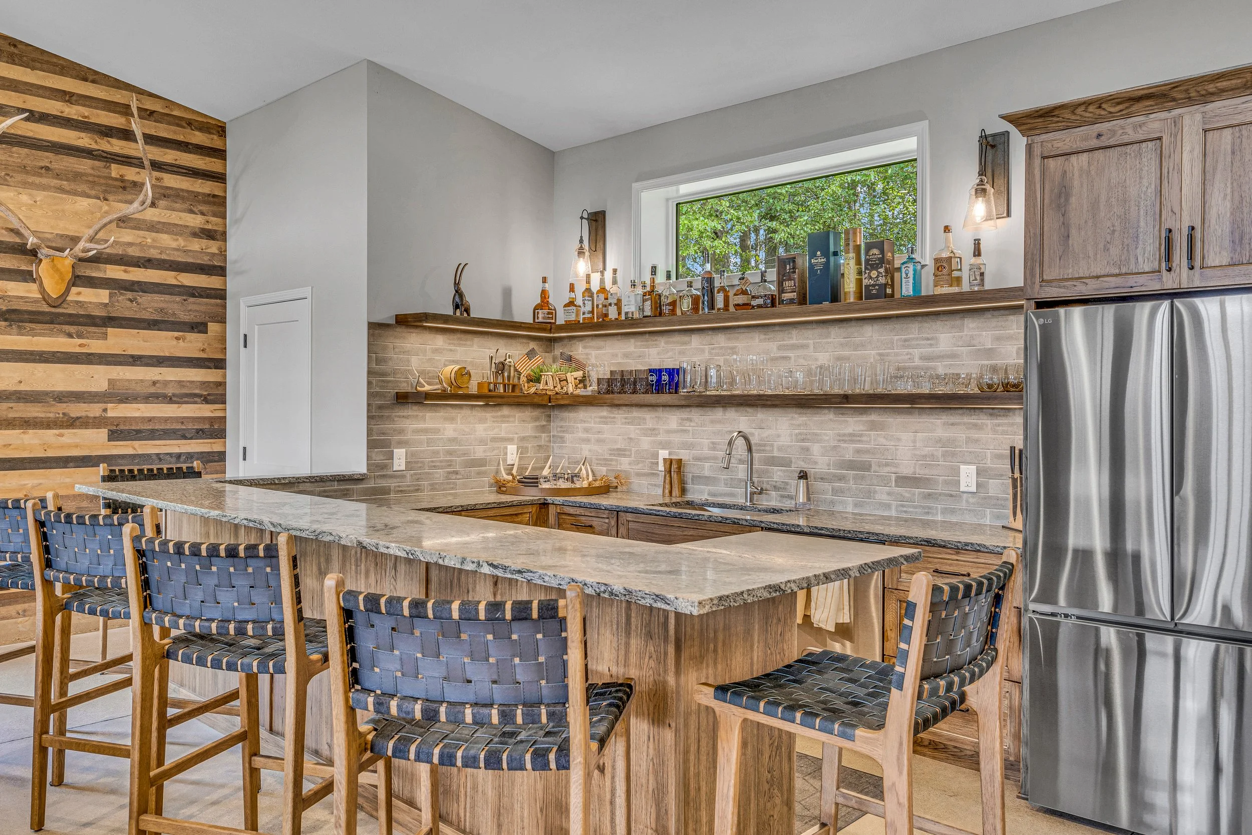 Modern kitchen with a wooden and stone island, open shelving with liquor bottles and glassware, a large window, and a stainless steel refrigerator.