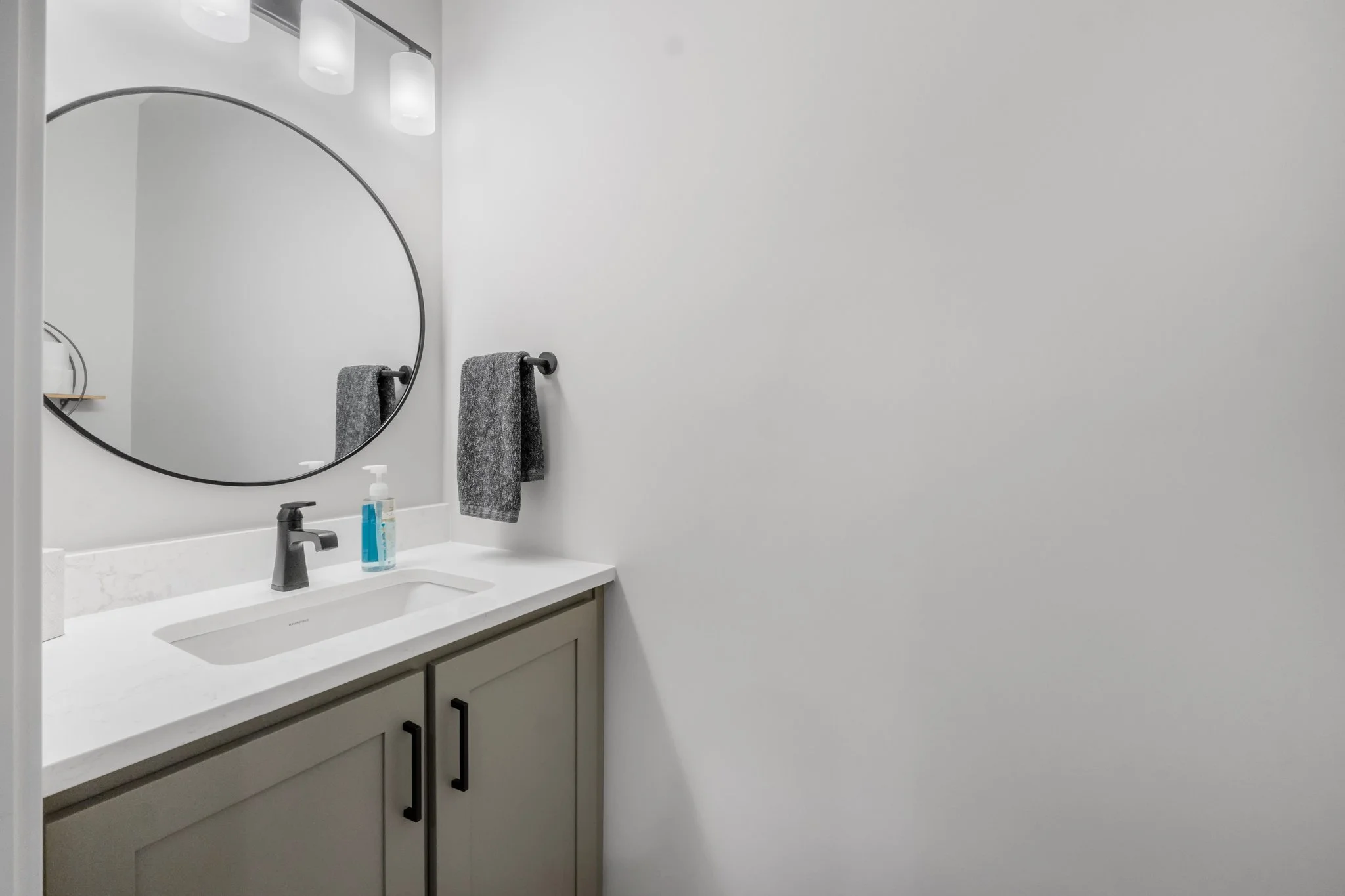 Bathroom sink with a round mirror, gray towels on a black towel bar, a soap dispenser on white countertop, and gray cabinets.