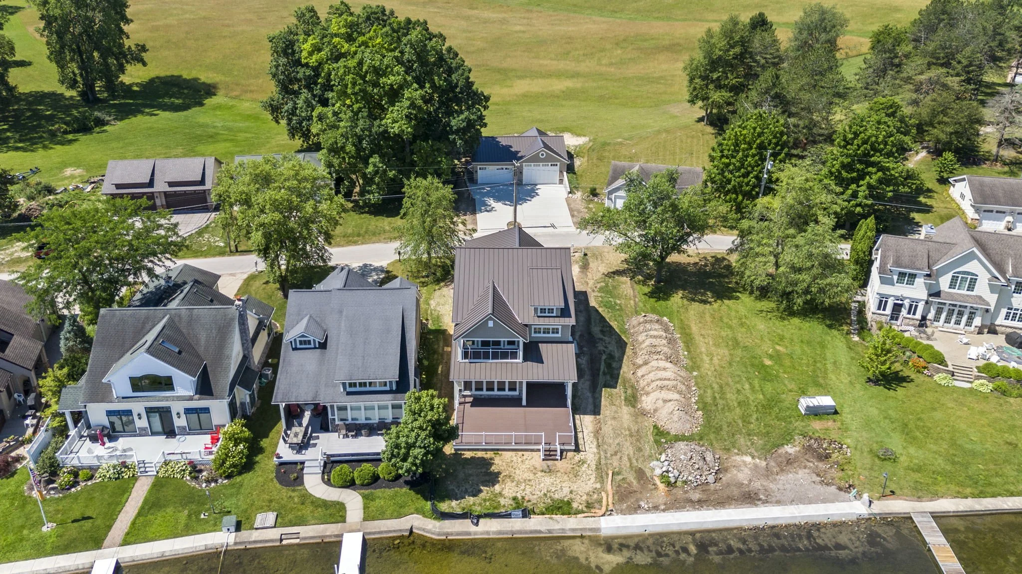 Aerial view of a neighborhood with several houses, trees, and a waterway at the bottom. One house is under construction, with dirt and construction materials nearby.