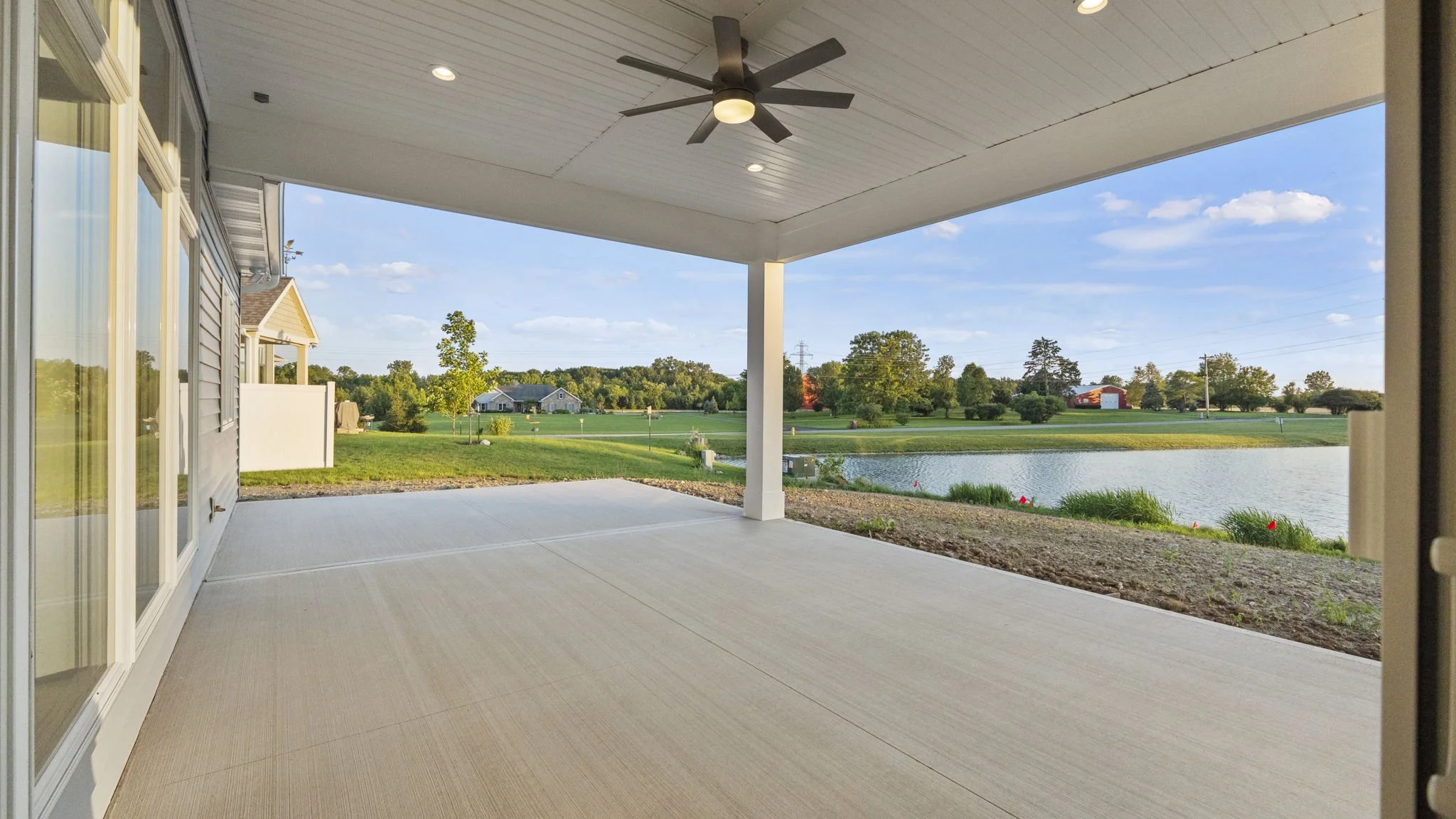 A covered patio overlooking a grassy yard, small pond, and trees with houses in the distance, under a blue sky with clouds.
