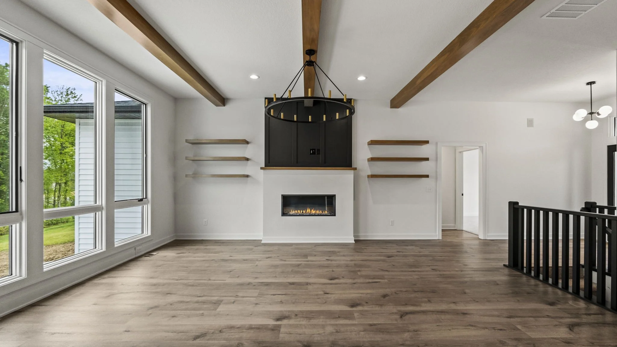 Empty living room with wooden floors, large windows, a fireplace, floating shelves, and black and wood accents.