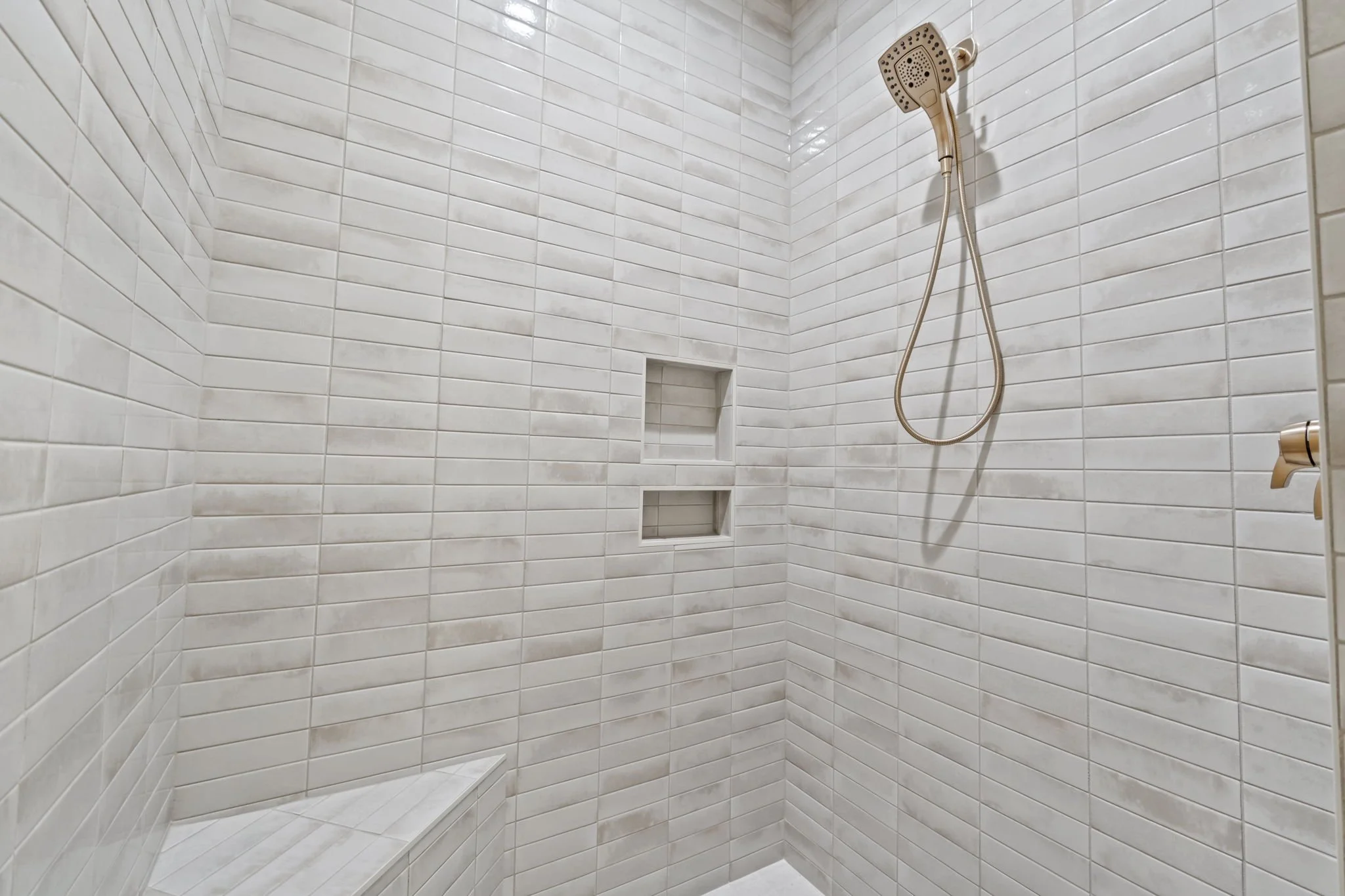 A tiled shower with beige subway tiles, a built-in shower seat, oval control knob, and a handheld showerhead mounted on the wall.
