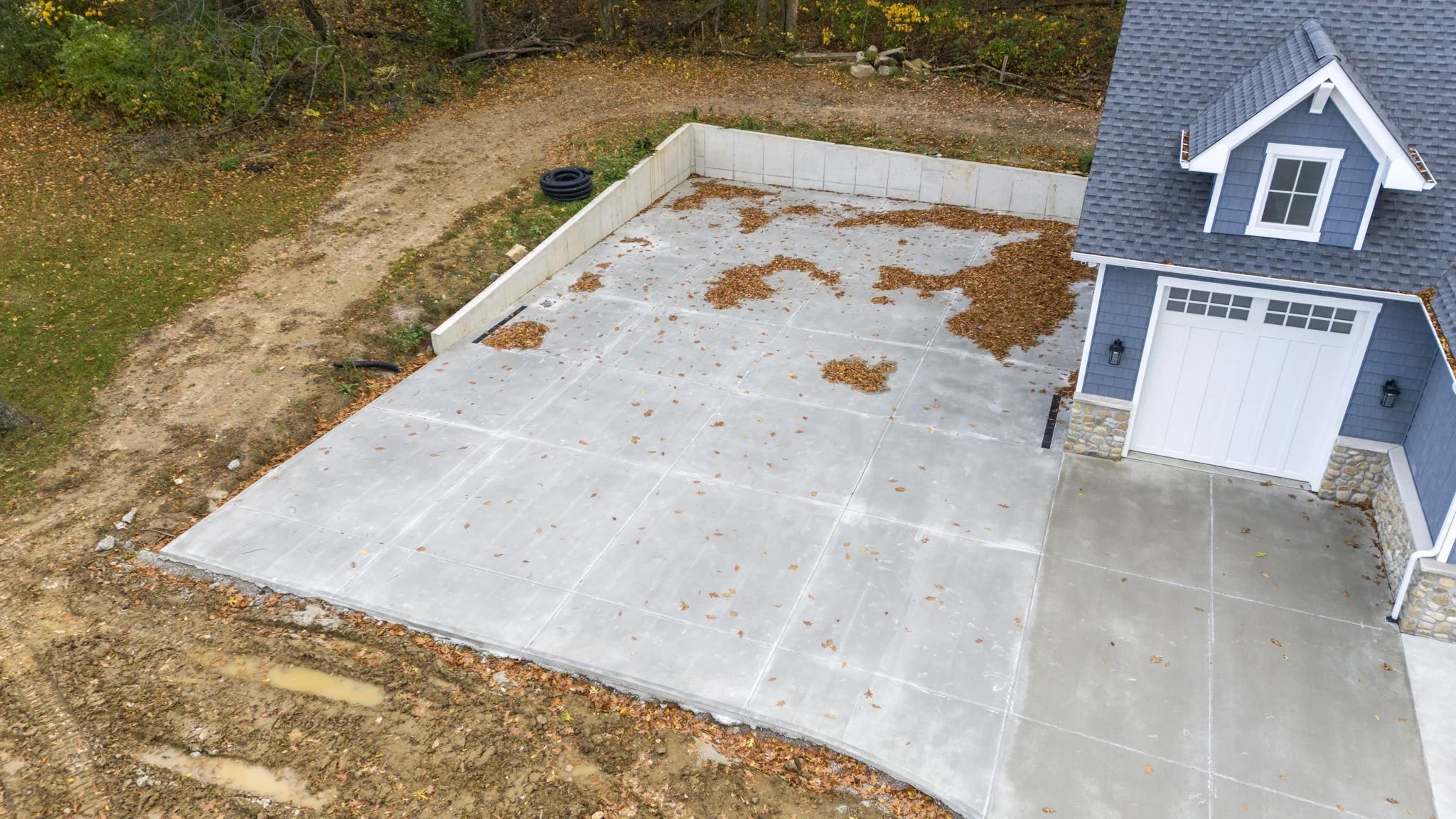 An aerial view of a newly poured concrete backyard patio with scattered leaves, adjacent to a blue house with white trim and mortar stone accents, surrounded by dirt and grass.