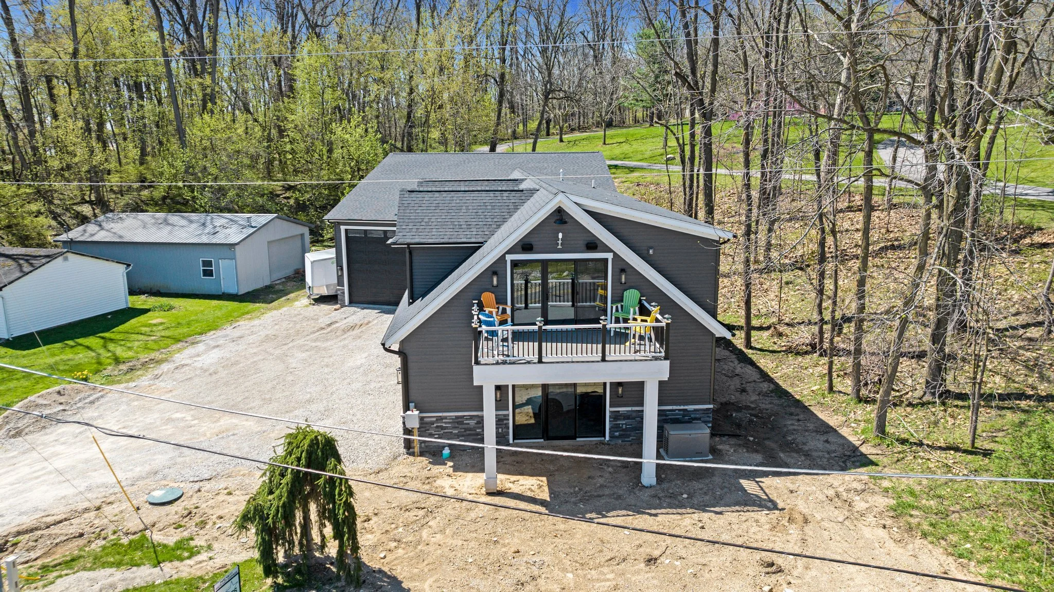 A two-story house with a balcony, gray exterior, and black roof, surrounded by trees and a backyard with a gravel driveway and green lawn.