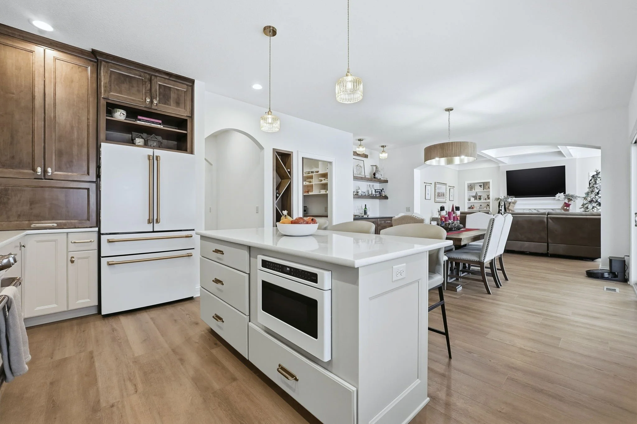 Open-concept kitchen and dining area with white and wood cabinetry, a white island with microwave, and a dining table with upholstered chairs. The background has a living room with a large flat-screen TV, sofas, and Christmas decorations.