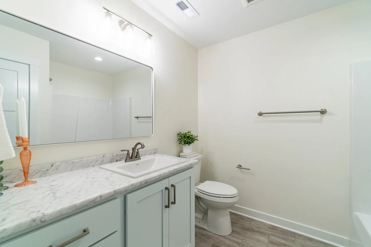 A clean, minimalist bathroom with a marble countertop, a white sink with a bronze faucet, a large mirror, and a white toilet. There are green plants and candles as decor, with towel bars and a wooden floor.