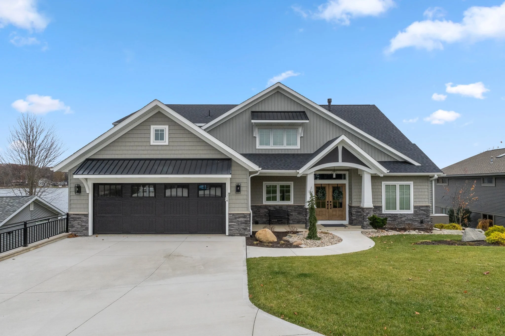 Modern two-story house with a gray exterior, black garage door, front porch, and landscaped yard under a blue sky.