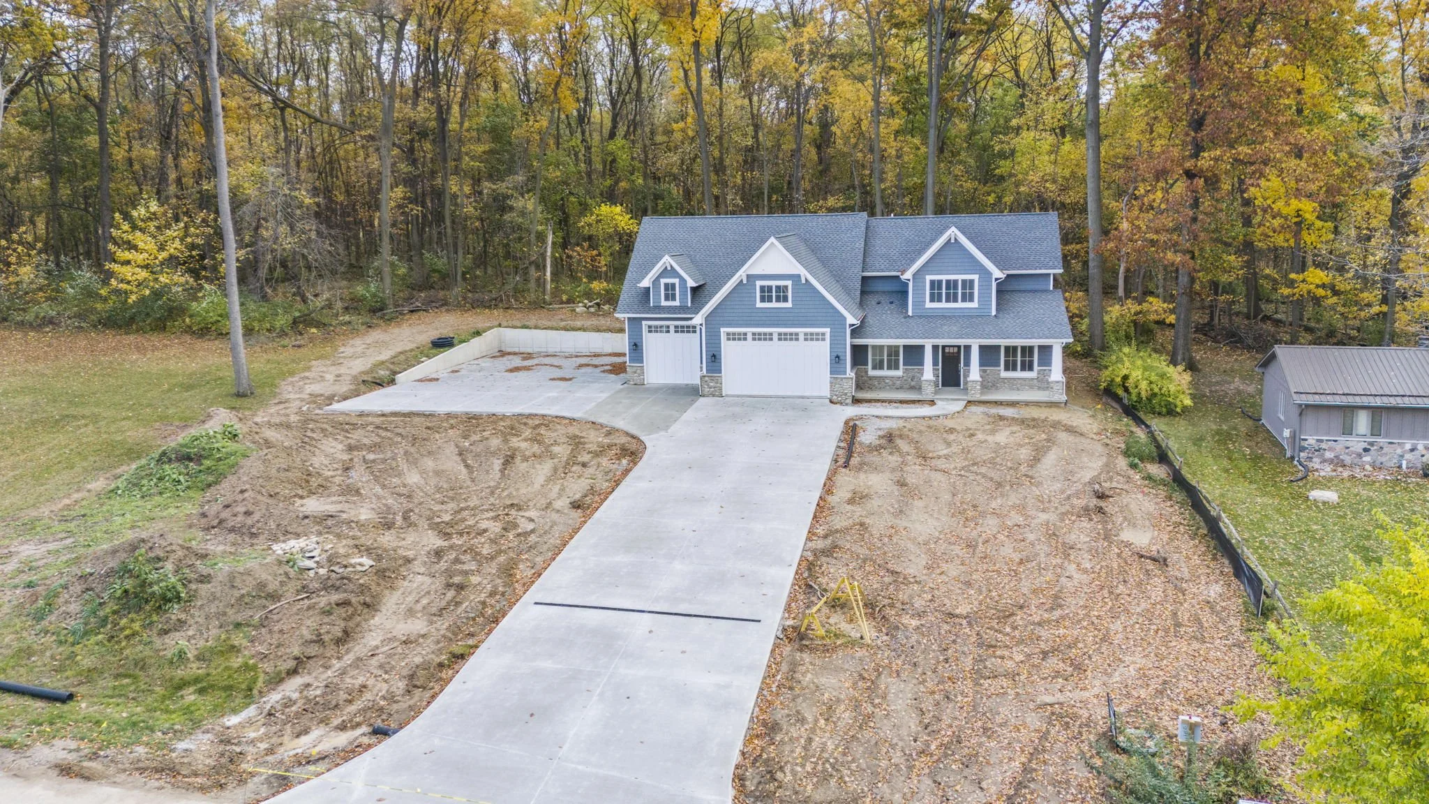 A newly constructed blue house with a large driveway, set in a partly wooded area with fall foliage.
