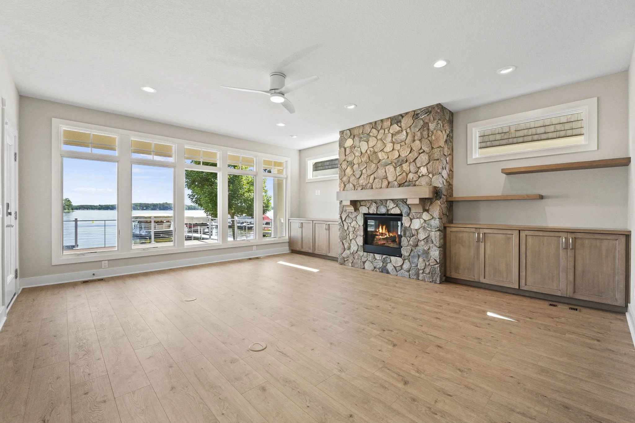 Empty living room with large windows overlooking a lake, stone fireplace, wooden built-in cabinets, shelves, and light wooden flooring.