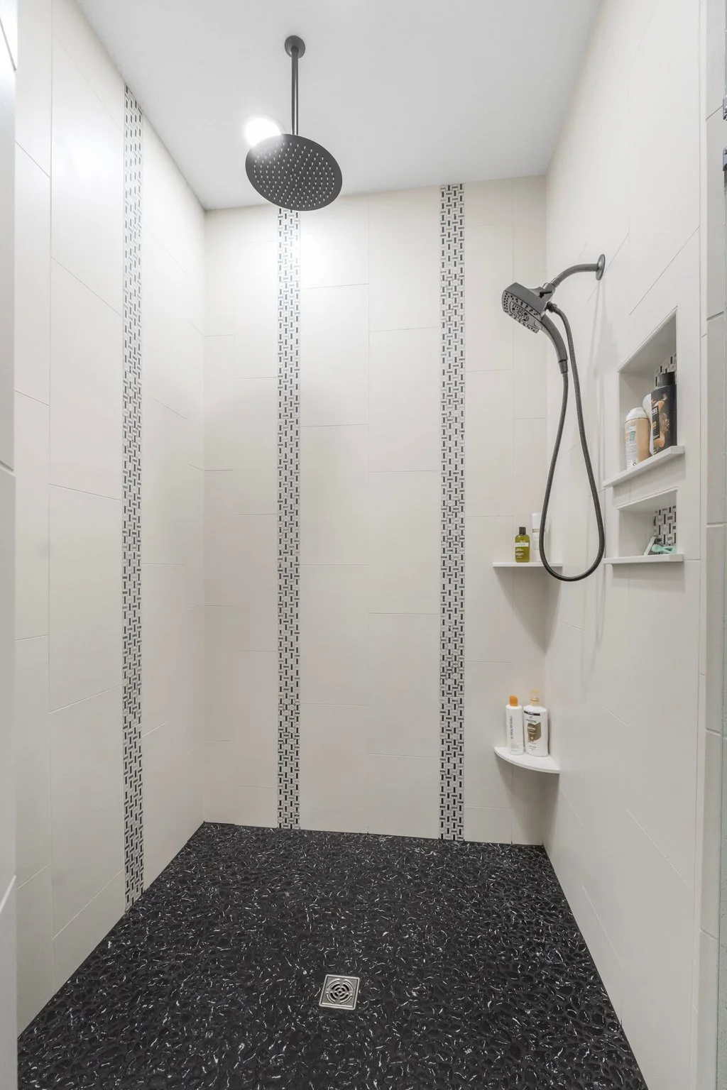 Modern shower with black and white patterned tiles, black rain showerhead, and built-in shelves with toiletries.