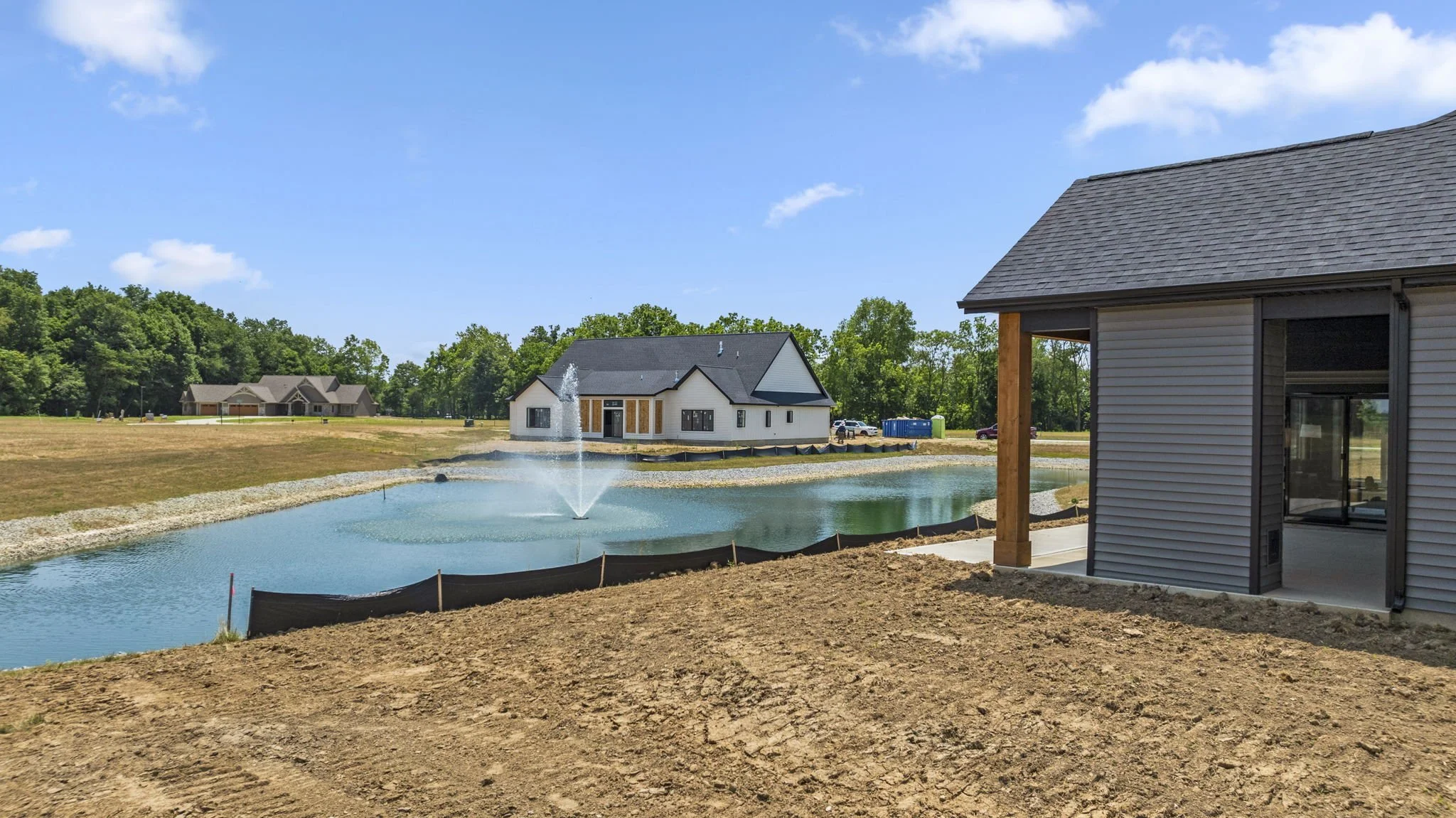 Under construction houses near a pond with a fountain, a house with white walls and black roof in the background, and a new house with gray siding and dark roof in the foreground, surrounded by dirt and green trees.