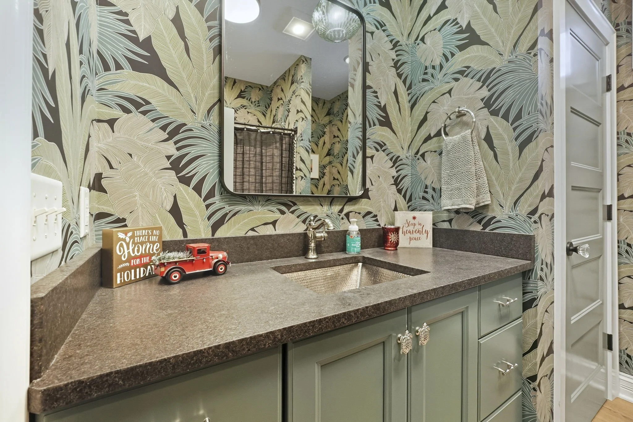 Bathroom vanity with a green cabinet, a brown countertop, and a mirror. The wall has a tropical leaf wallpaper. Decor items include a small red vintage truck, a sign, hand soap, a red candle holder, and a small decorative sign with holiday messages. 