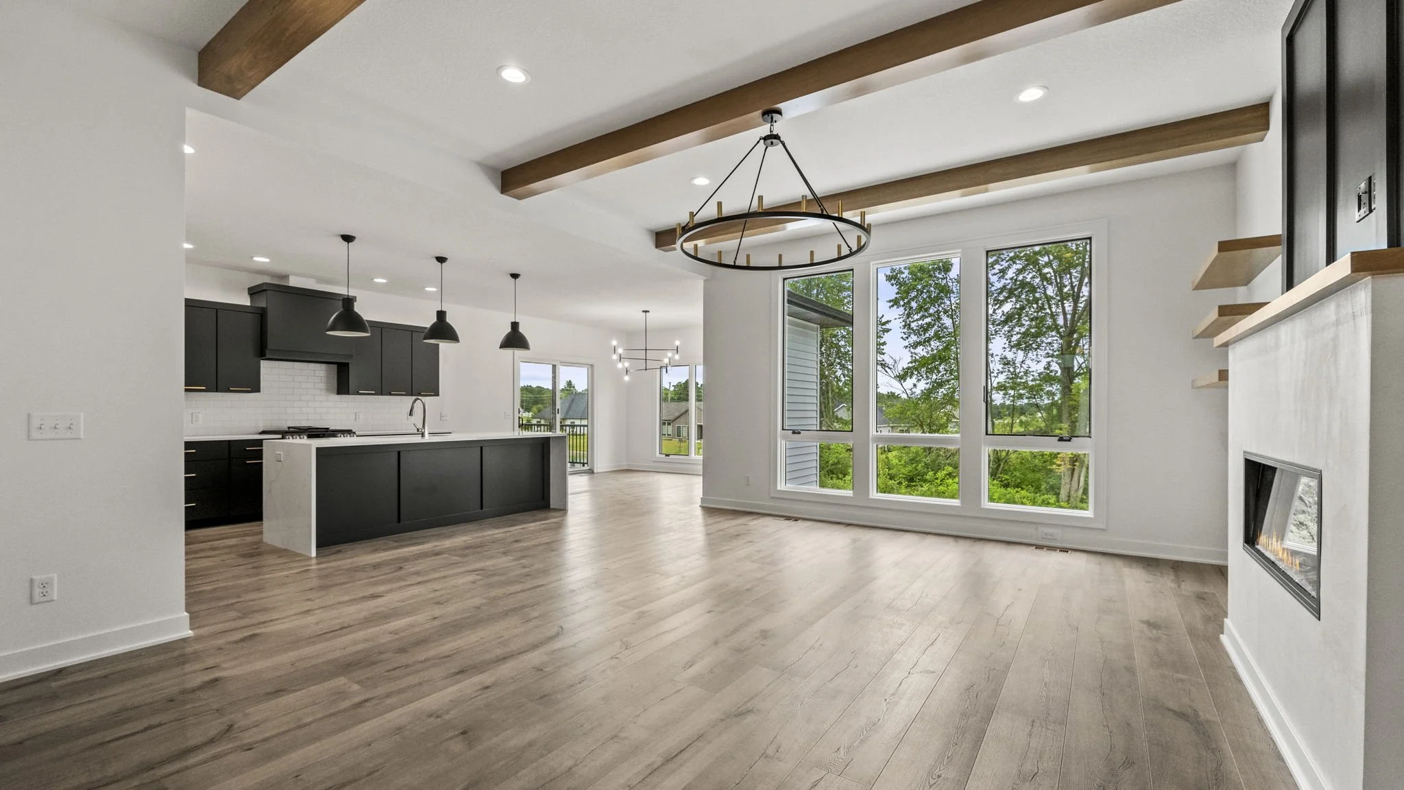 Open-concept living space with large windows showing green trees outside, a modern kitchen with black cabinets and pendant lights, light wood flooring, and a fireplace with shelves built into the wall.
