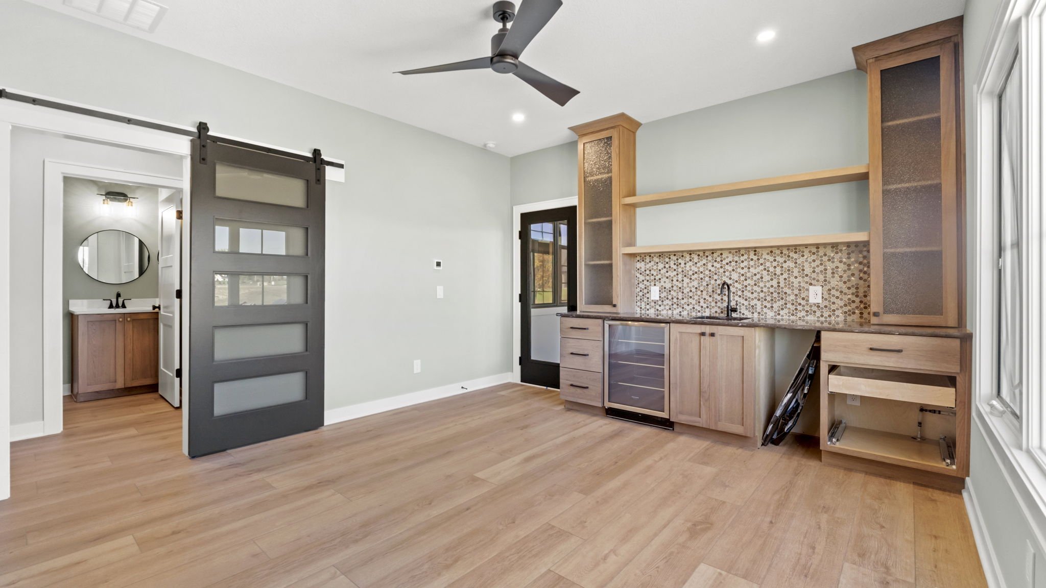 Empty room with light green walls, a ceiling fan, wood flooring, a built-in cabinet with open shelves, a small wine cooler, and a sliding barn door leading to a bathroom with a vanity and round mirror.