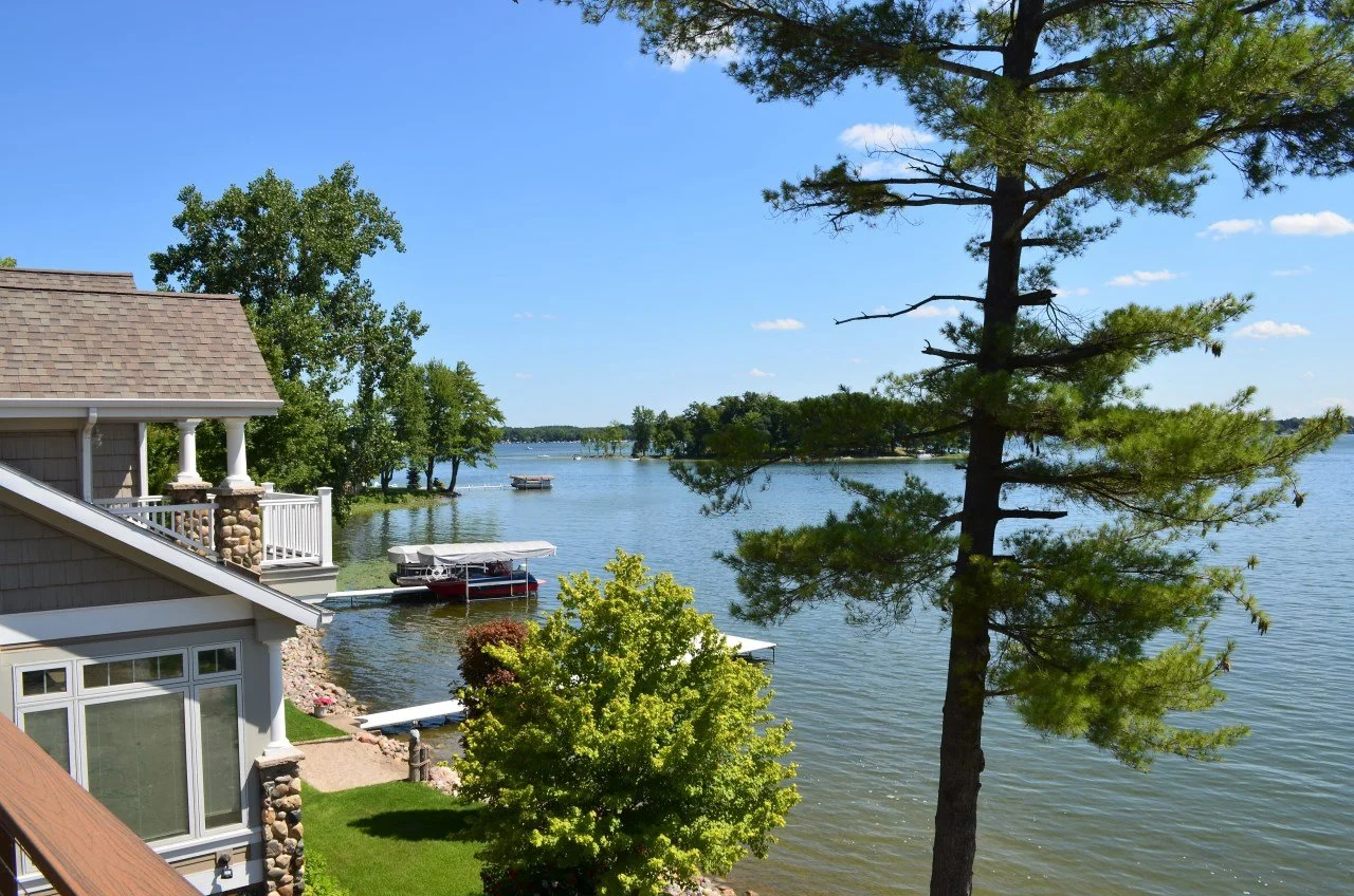 A lakeside view showing part of a house with a sloped roof, a dock with boats, a large tree in the foreground, and a calm lake with distant trees on the shoreline under a clear blue sky.