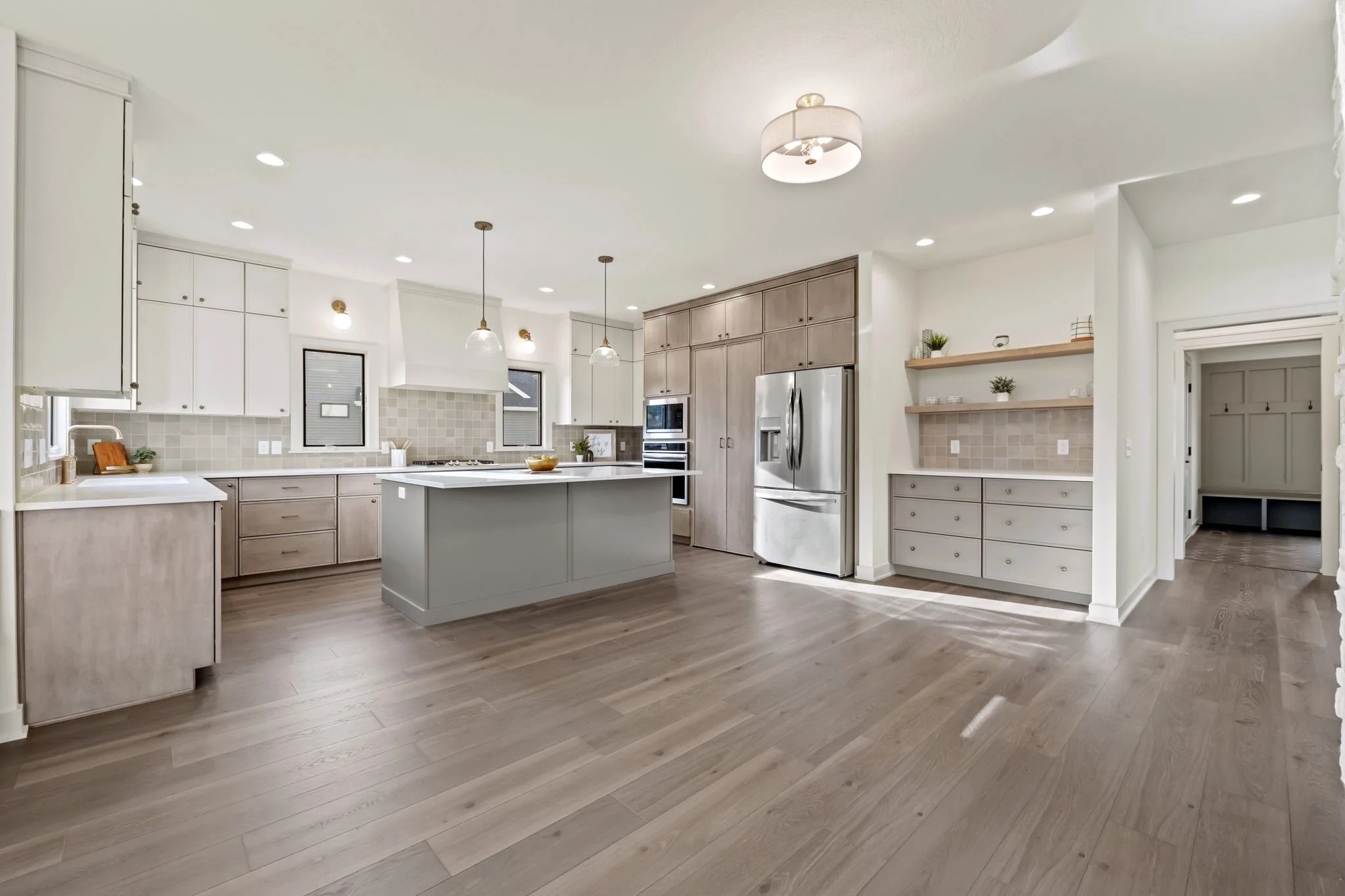 Modern kitchen with white and light wood cabinets, stainless steel appliances, and a large central island with a white countertop. There are pendant lights, recessed lighting, and a tiled backsplash. The floor is wood, and there is a small nook with 