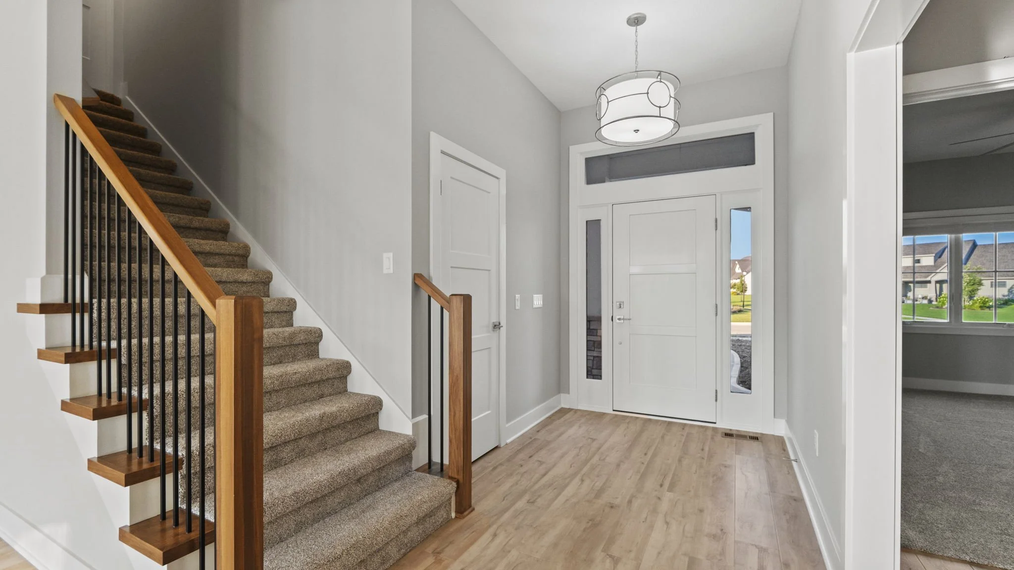 Interior of a modern house entryway with a staircase, front door, and a view into a living room with large window.