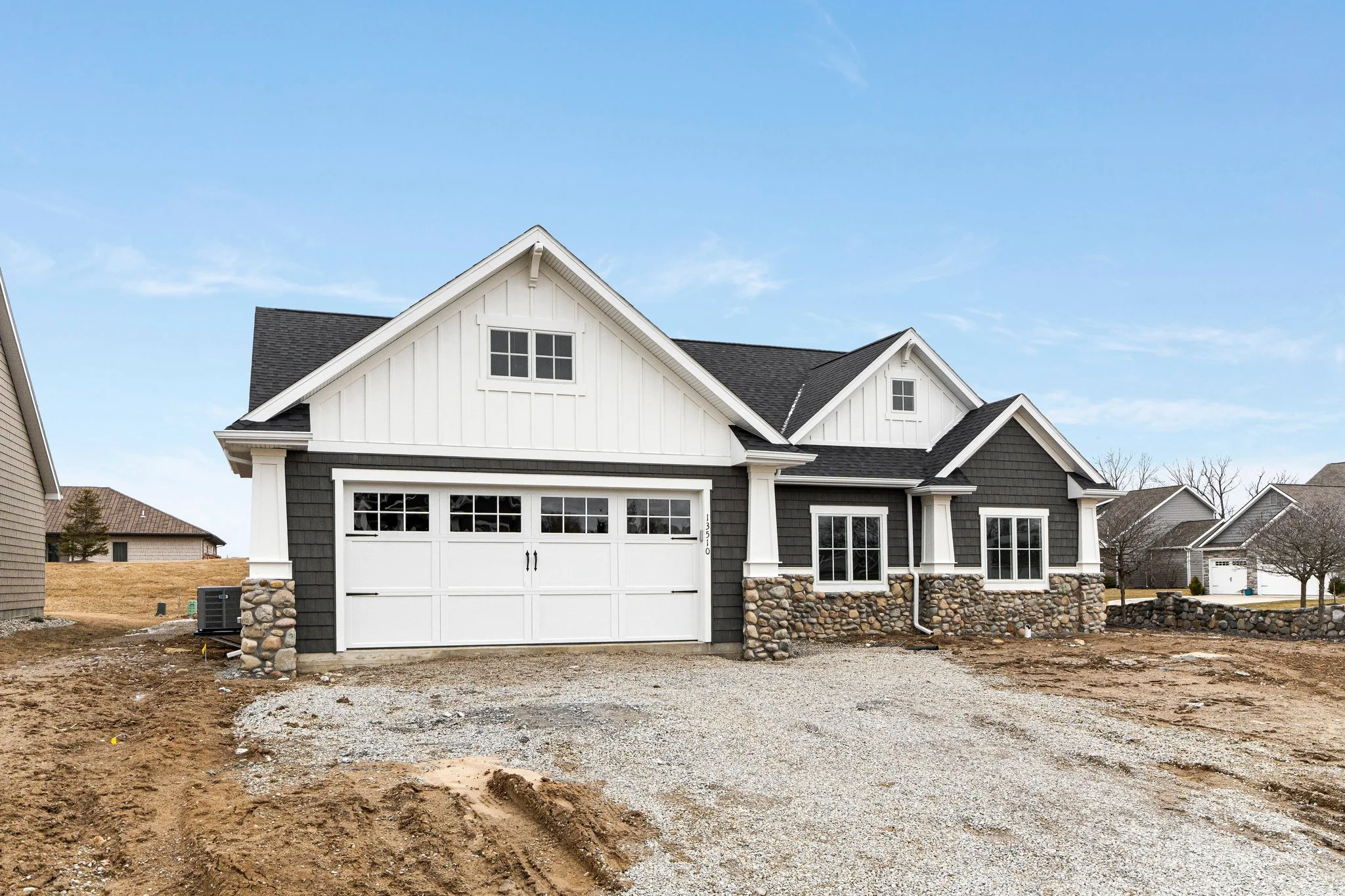 Newly constructed suburban house with gray and white exterior, stone accents, and a white garage door, on a gravel driveway with adjacent houses and trees.