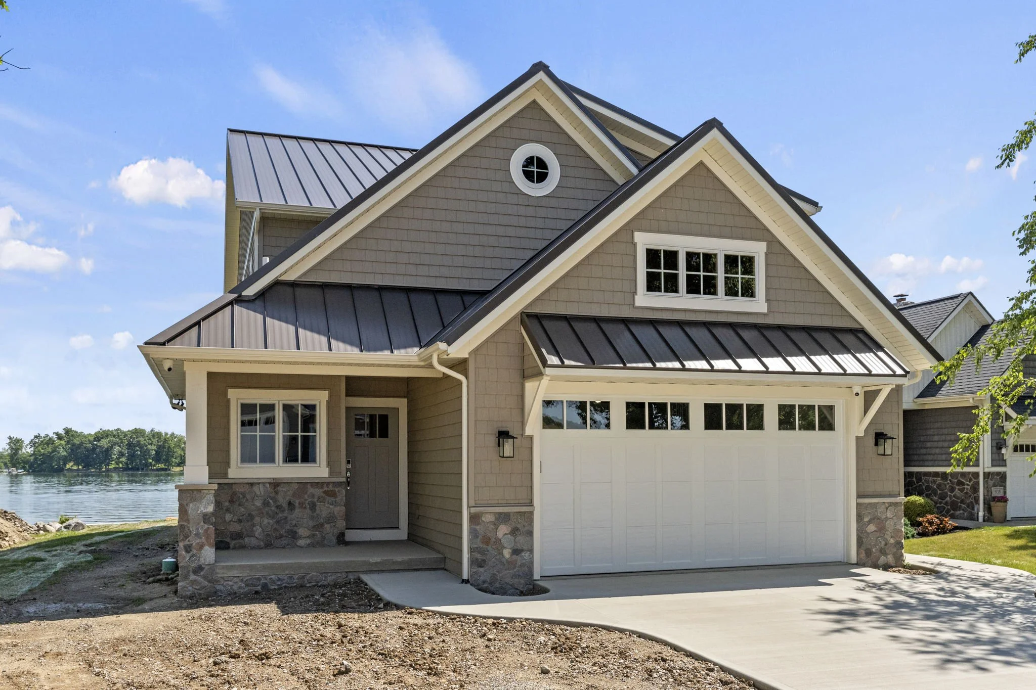 A two-story house with beige siding, stone accents at the base, and a metal roof, situated near a body of water, with a concrete driveway leading to a garage.