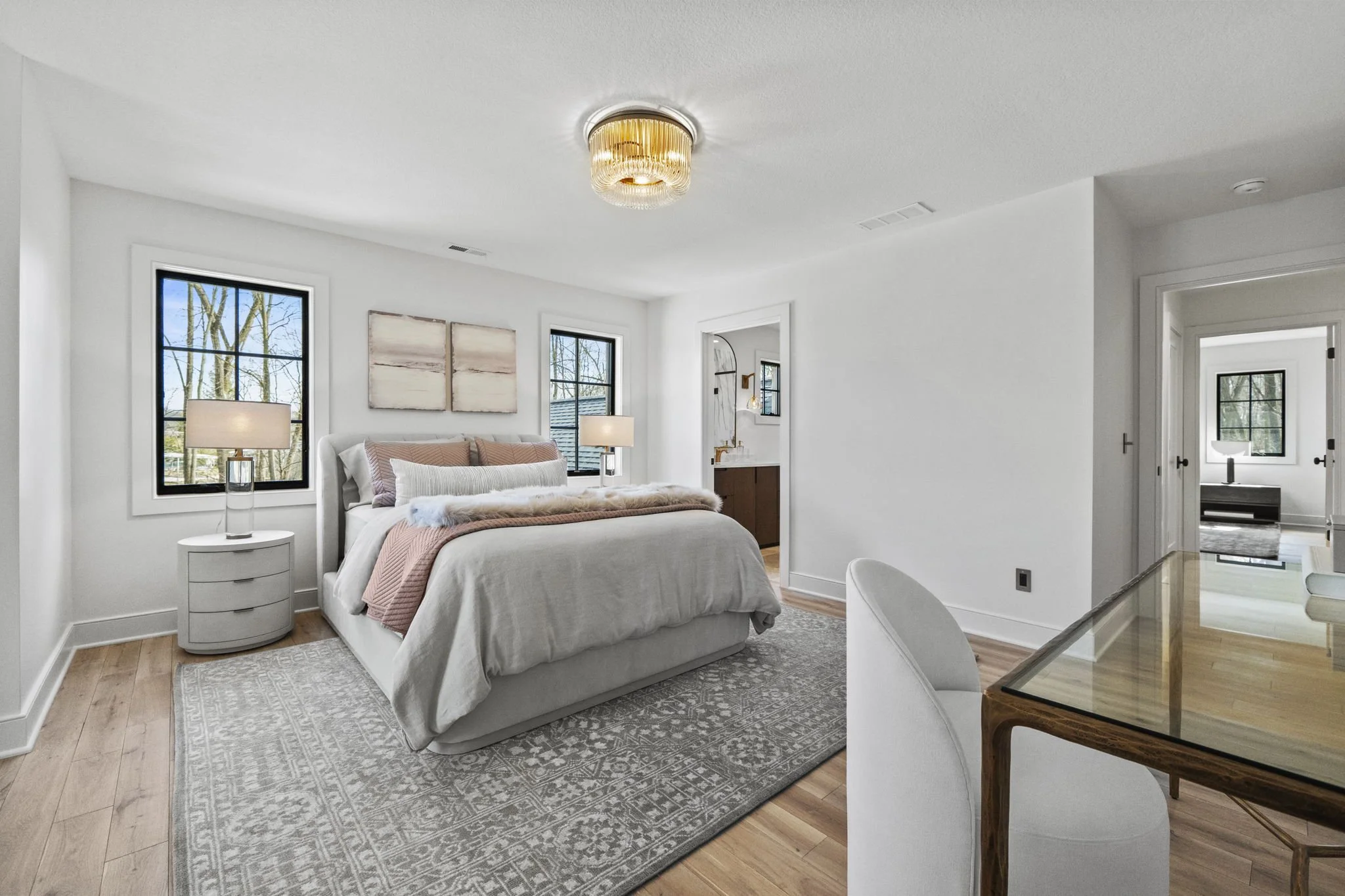 Modern bedroom with white walls, wood flooring, and a large bed with neutral and pink bedding. There are two windows behind the bed, two nightstands with lamps, a rug under the bed, and a chandelier on the ceiling. A view through an open doorway show