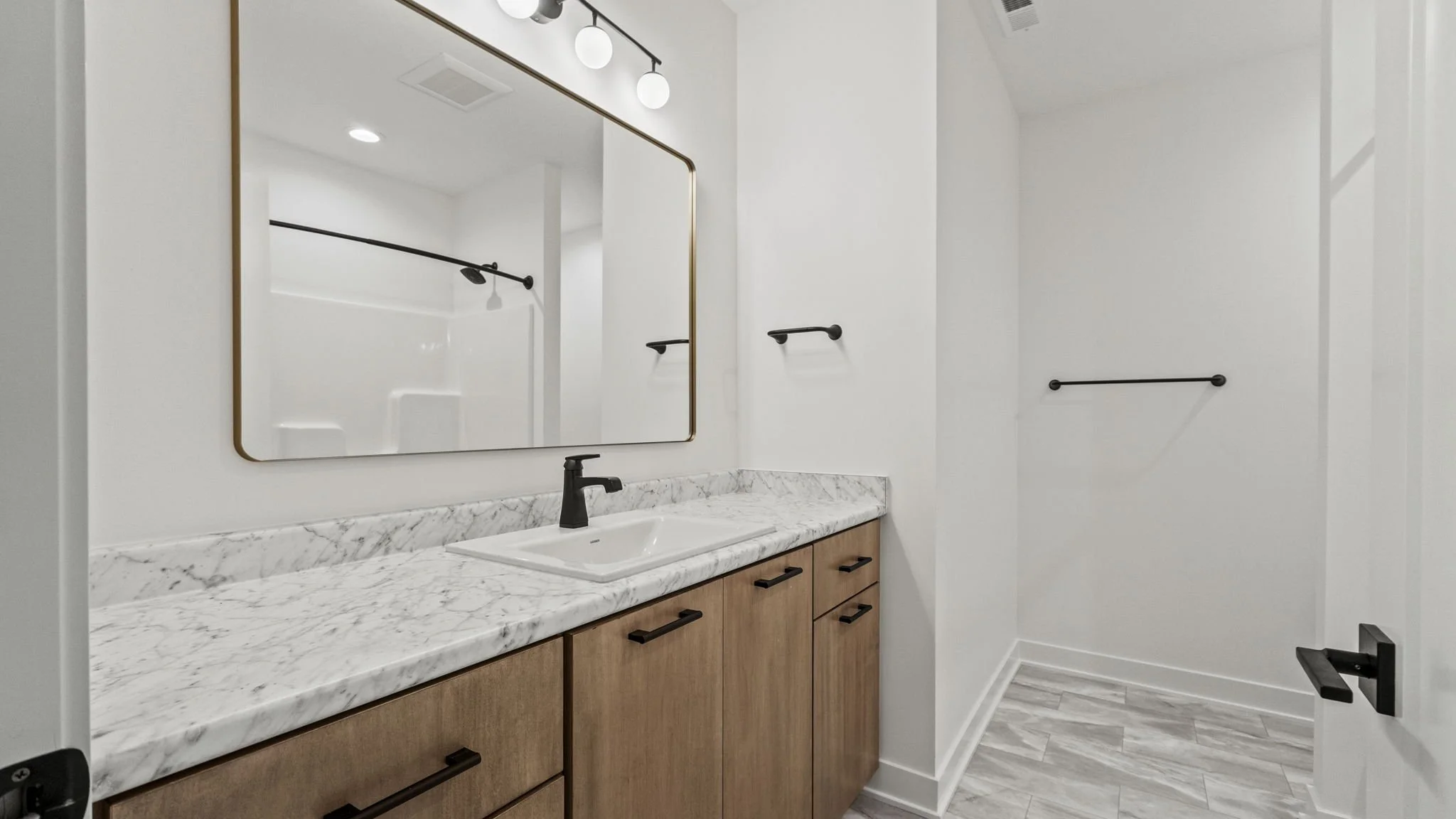 Bathroom with white walls, a marble countertop with a sink, wooden cabinets, a large mirror with a gold frame, black fixtures, and black towel bars. The floor has light-colored tiles.