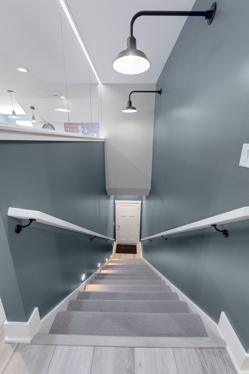 A staircase descending from a second-floor landing with gray walls, white handrails, and built-in wall lighting. At the bottom, a closed white door is visible.