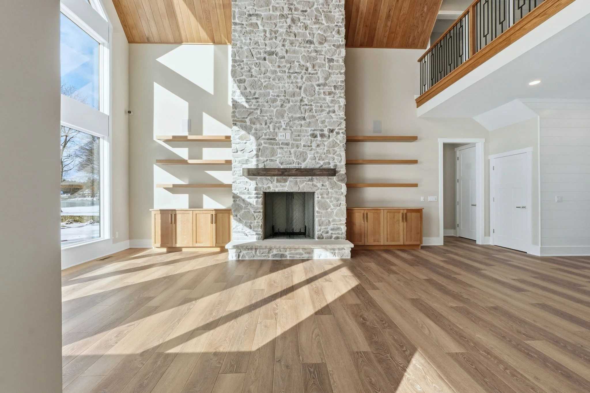 Empty living room with large windows, a stone fireplace with a wood mantel, built-in wooden cabinets, and floating wooden shelves, with hardwood floors and a high wooden ceiling.