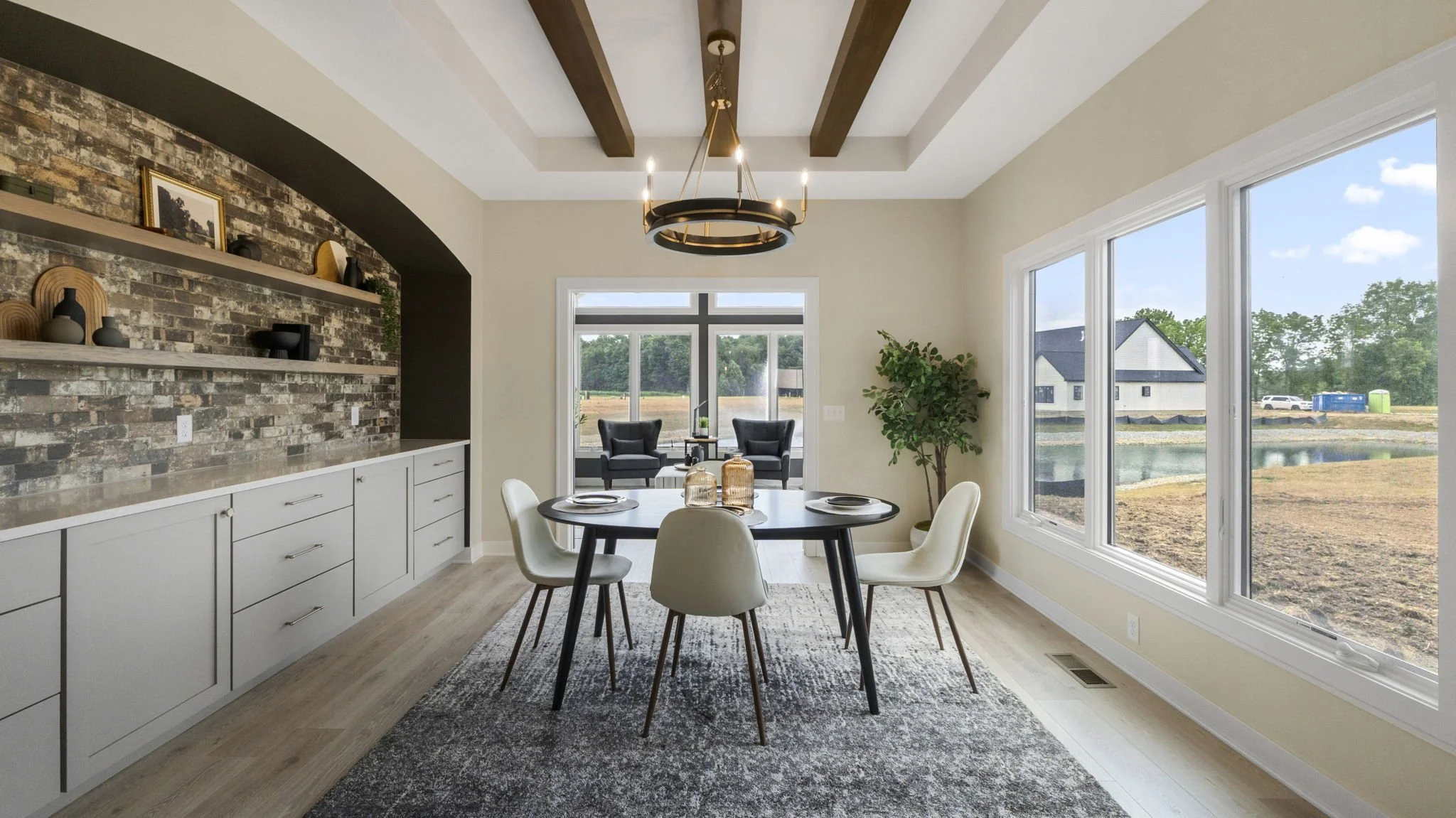 Modern dining room with a round black table and six white chairs, large windows showing a yard and pond outside, built-in sideboard, decorative shelves with vases, and a chandelier.