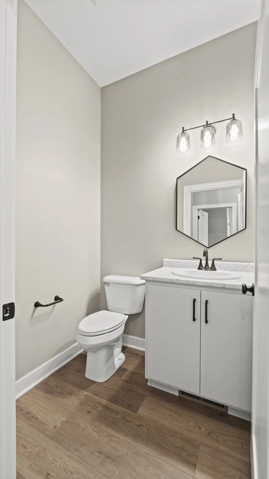 A small bathroom with beige walls, a white toilet, a white vanity with a marble countertop, a hexagonal mirror, and a three-bulb light fixture.