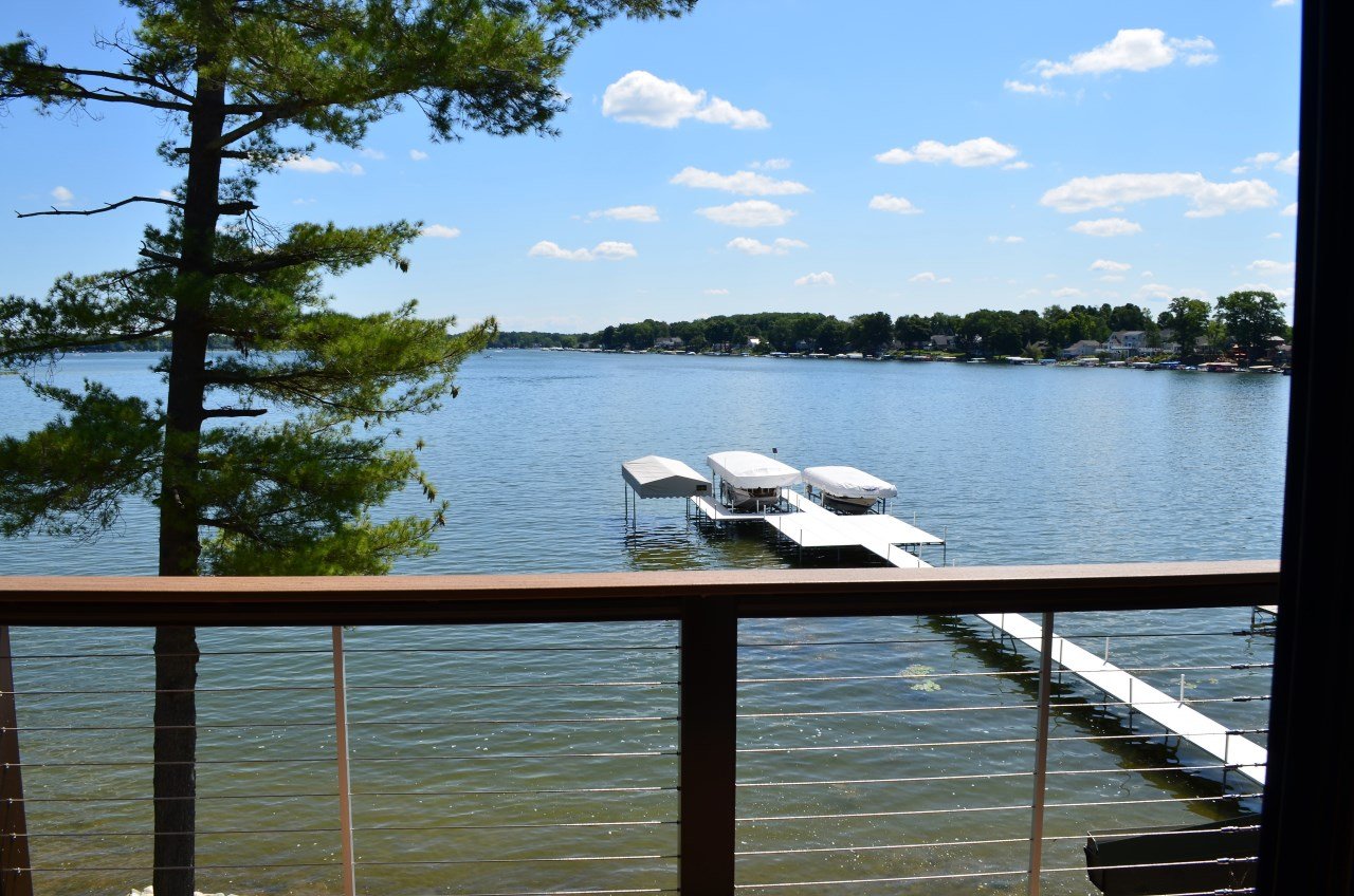 View of a lake from a balcony, with a tree on the left, a dock with boats under covers in the center, and houses along the shoreline in the distance.
