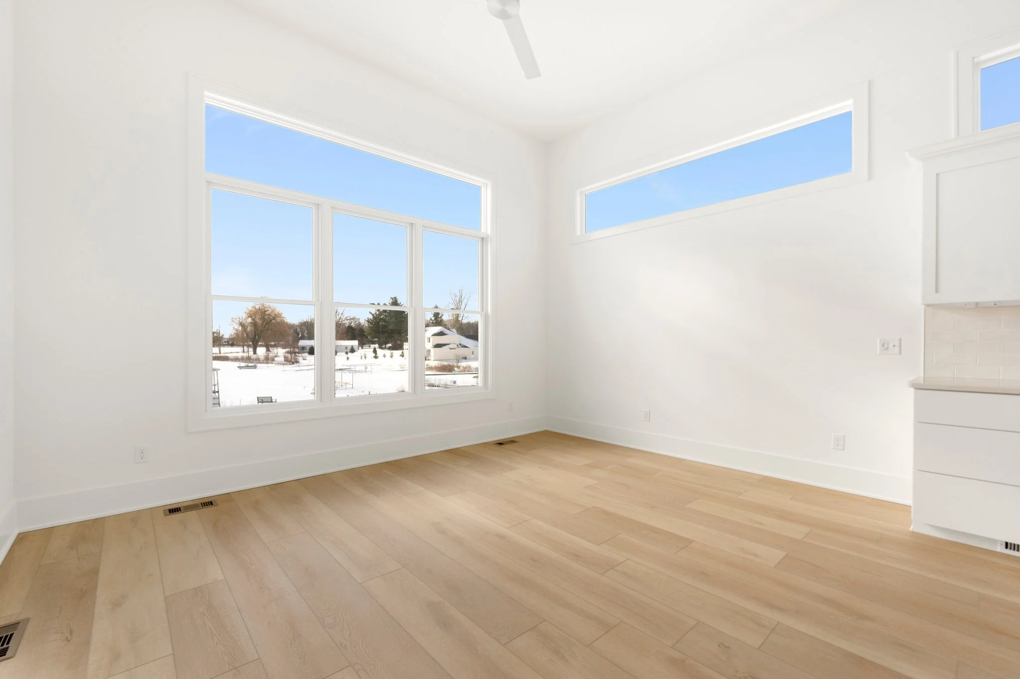 Bright empty room with large windows, white walls, wooden flooring, and a ceiling fan, with outdoor snow-covered landscape visible through the windows.