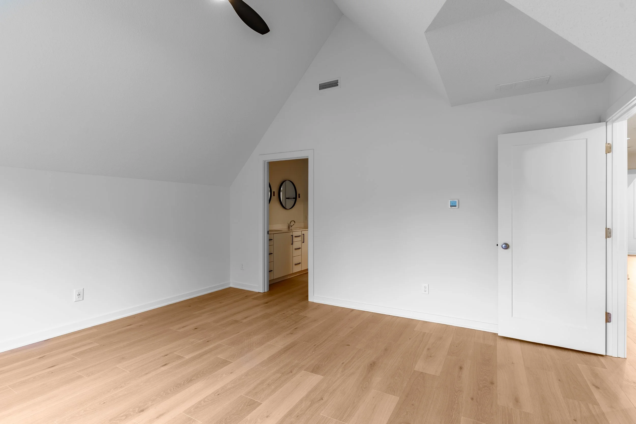 Empty room with white walls, a small door leading to a bathroom with mirrors, and a wooden floor.