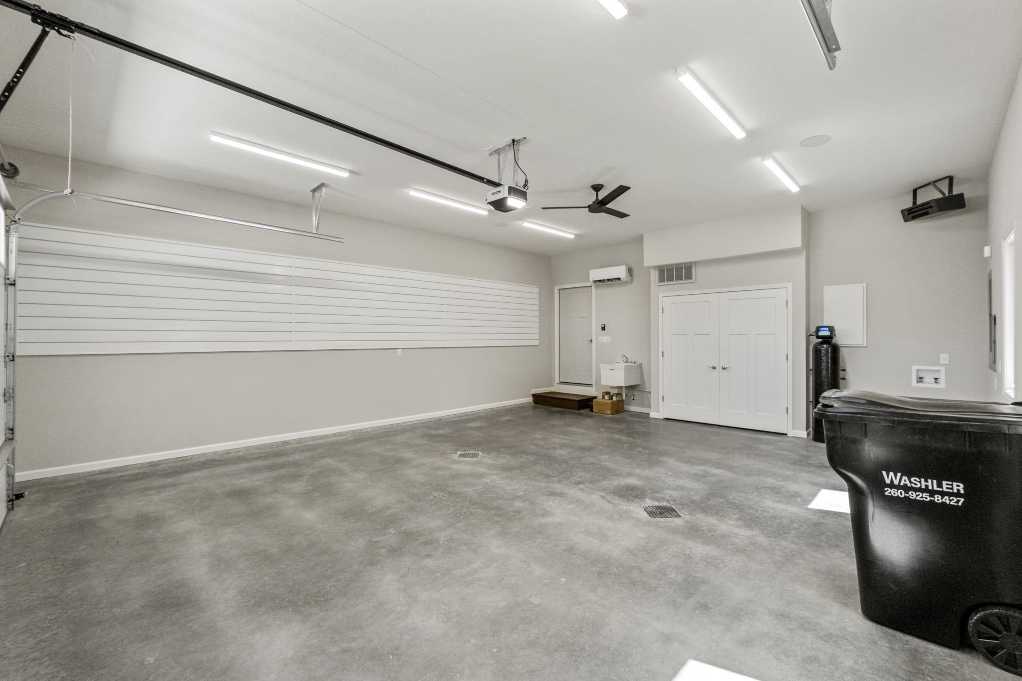 Empty garage with concrete floor, white walls, fluorescent ceiling lights, garage door, wall-mounted storage slat, small sink, black waste bin, and an exercise fan.