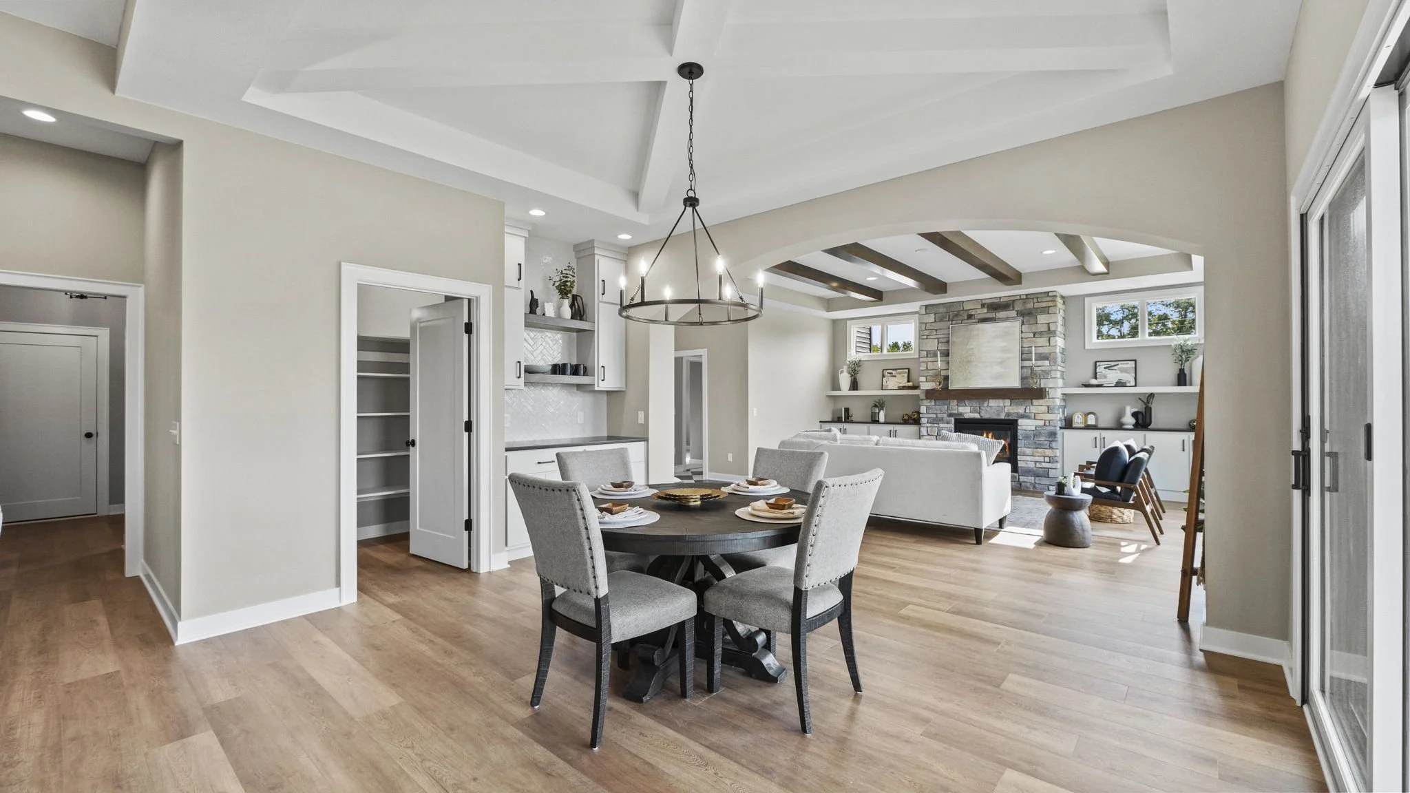 Open-concept living and dining area with hardwood floors, a round dining table with six chairs, a sofa facing a fireplace, shelves with decor, and large windows allowing natural light.